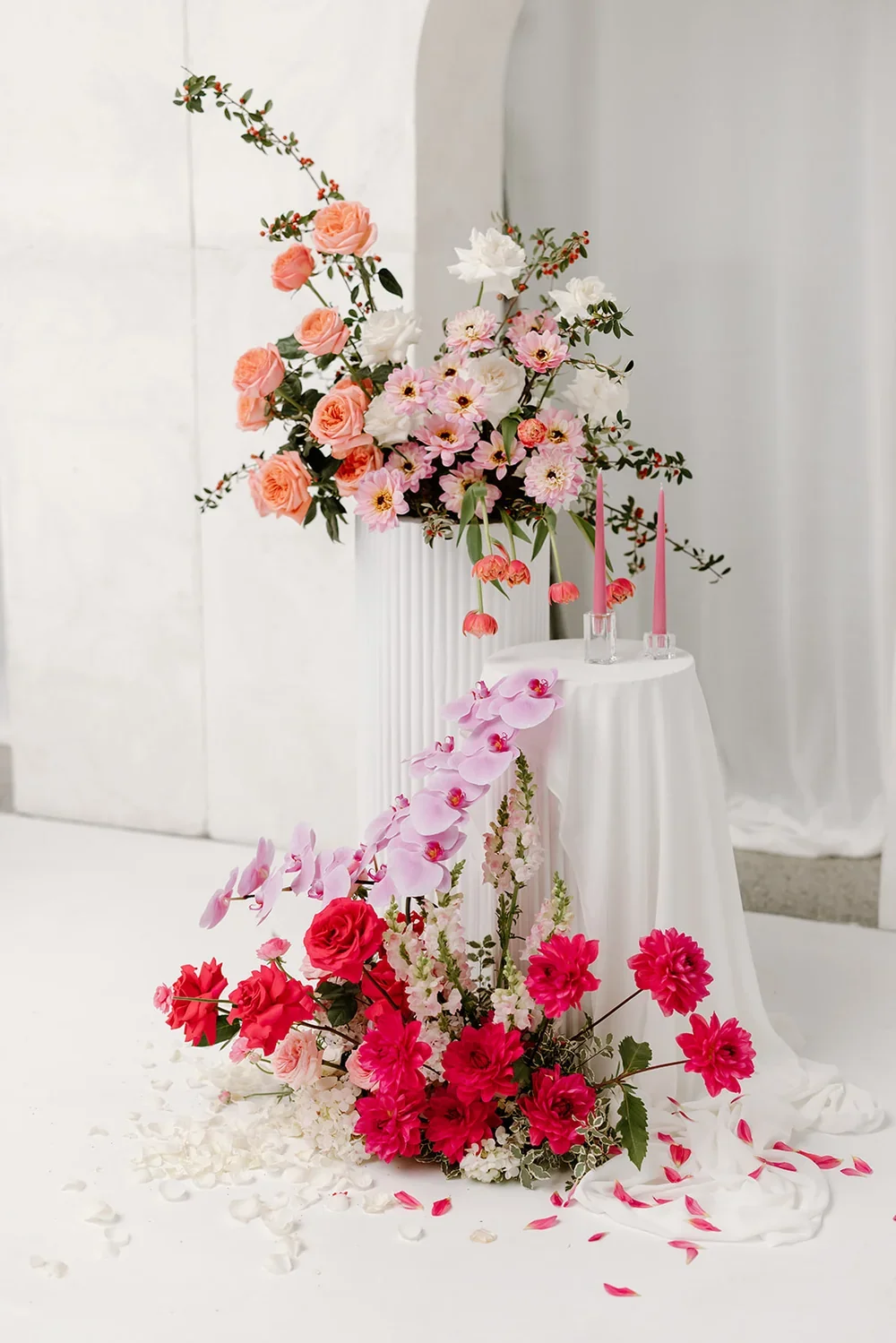 Two brightly coloured floral arrangements with pink roses, orchids and tulips clustered together at different heights with draped plinth and taper candles