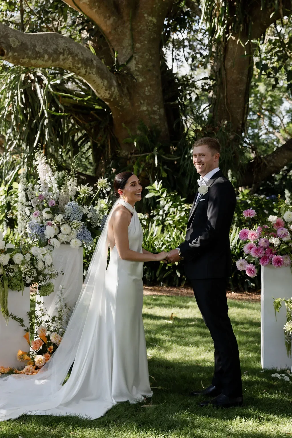 Bride and groom holding hands during an outdoor wedding ceremony beneath a large fig tree, surrounded by lush garden florals and white floral pillars.