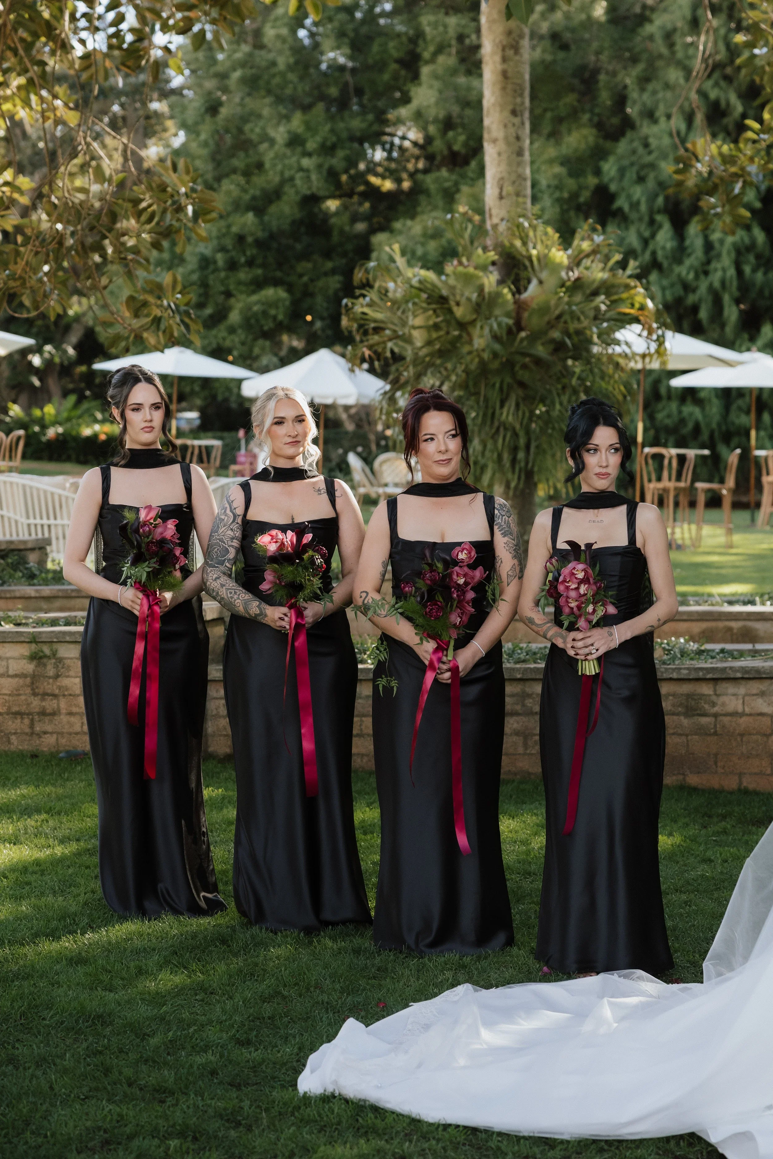 Bridesmaids in black gowns holding deep red and pink bouquets with trailing ribbons on the lawn at Gabbinbar
