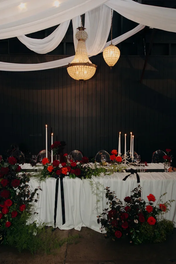 Red rose floral arrangement with white layered table linen, mirror runners, candlelight and vintage chandeliers hanging from the ceiling.
