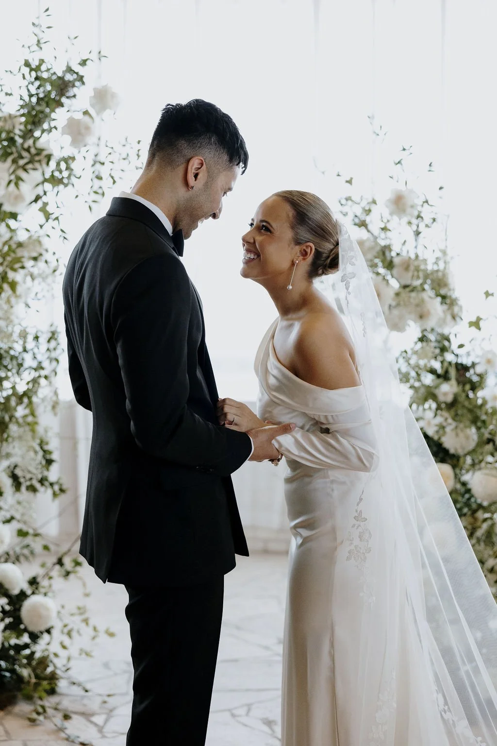 Bride and groom smiling and holding hands during intimate wedding ceremony surrounded by white florals.