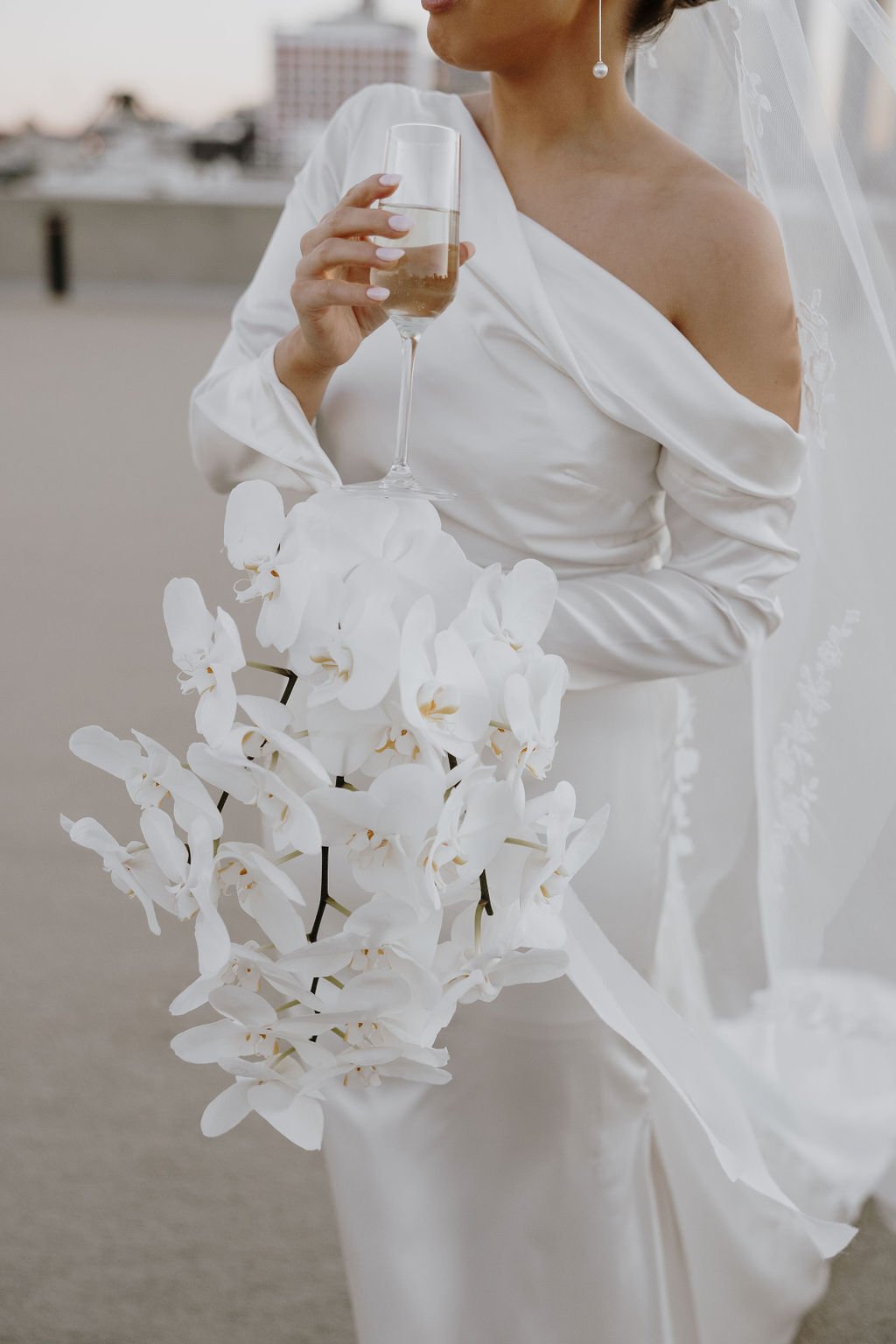 Bride holding champagne glass and white orchid bouquet, showing off elegant off-shoulder silk gown.”