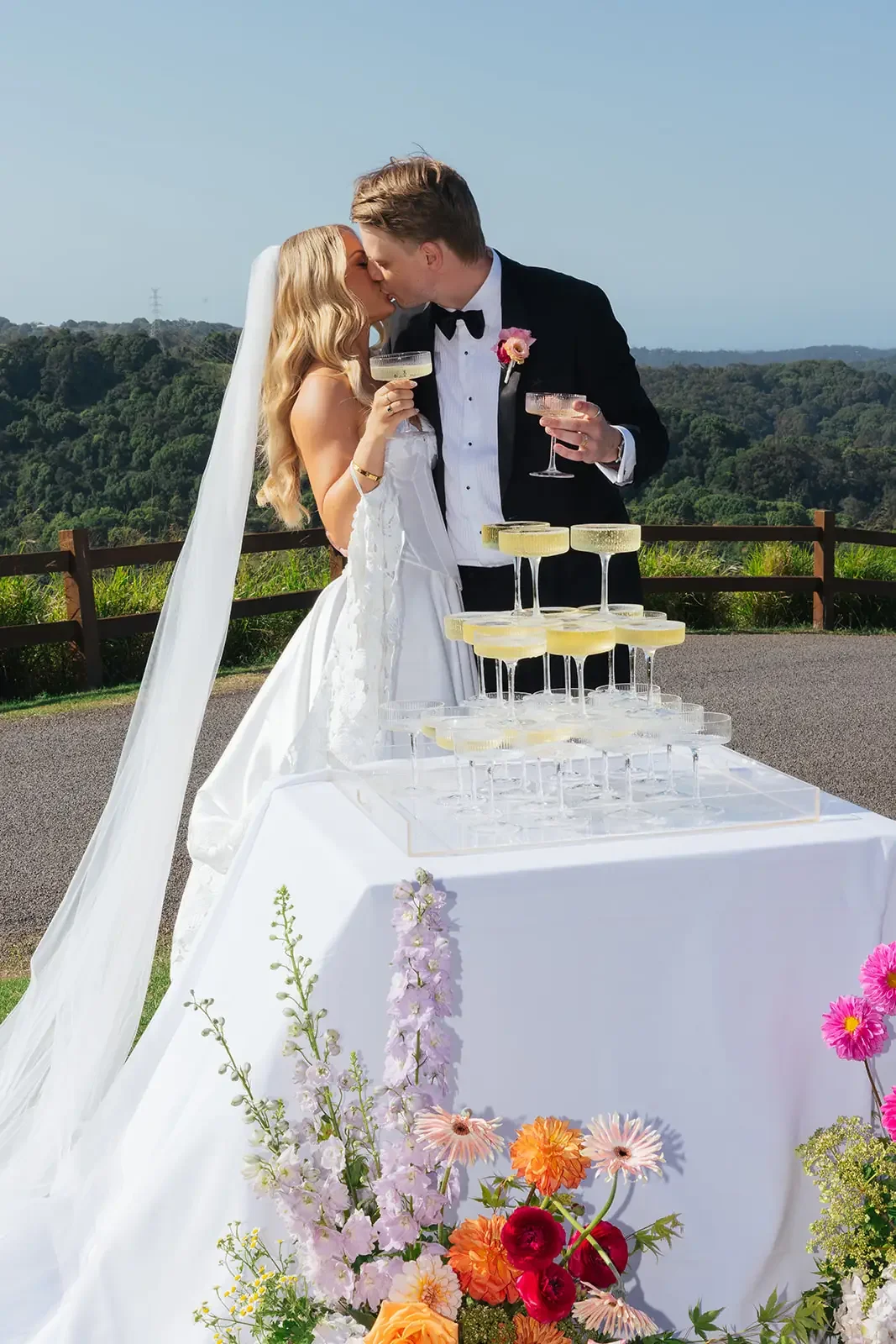 Bride and groom kissing beside a champagne tower, surrounded by bright garden-style floral arrangements in pink, orange and pastel tones.