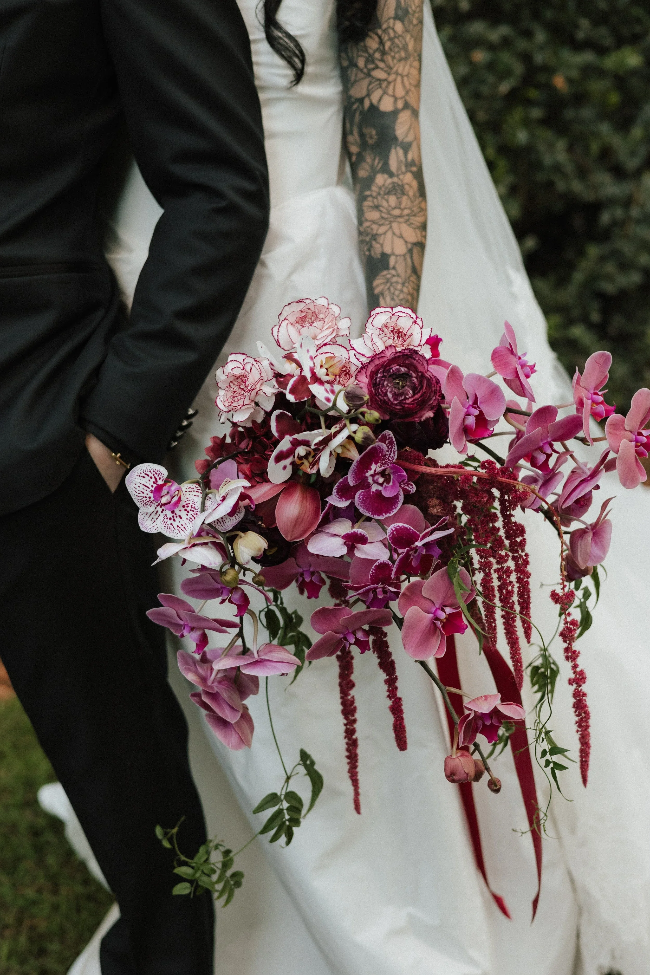 Trailing bridal bouquet of pink orchids and deep red blooms with long red ribbons against a white gown