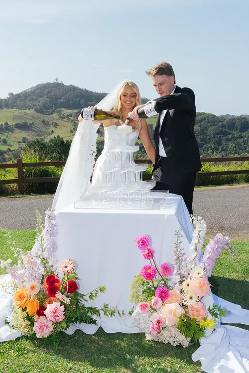 Bride and groom pouring champagne into a glass tower styled with bright floral arrangements