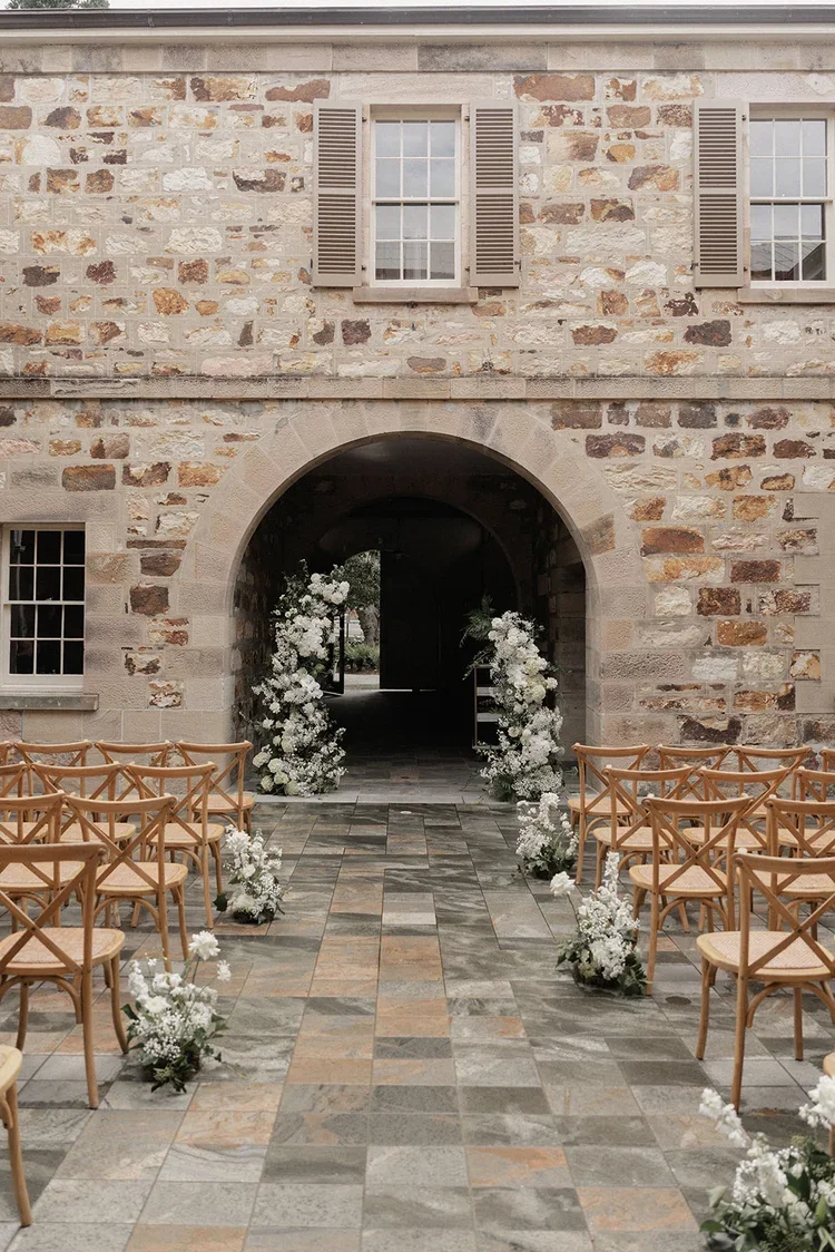 Ceremony setup at a historic stone venue with wooden chairs and white floral arrangements leading toward an arched entryway.