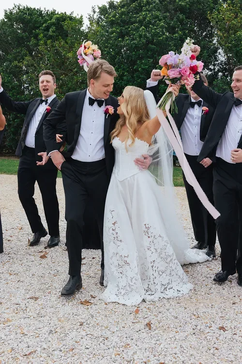 Bride and groom celebrating with their wedding party, holding a bouquet and smiling outdoors