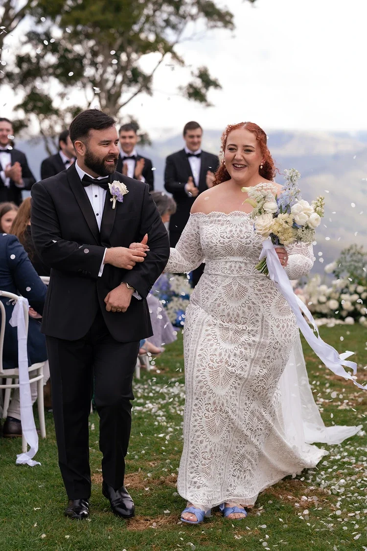 Bride and groom walking back up the aisle smiling as guests throw white petals during the ceremony recessional.