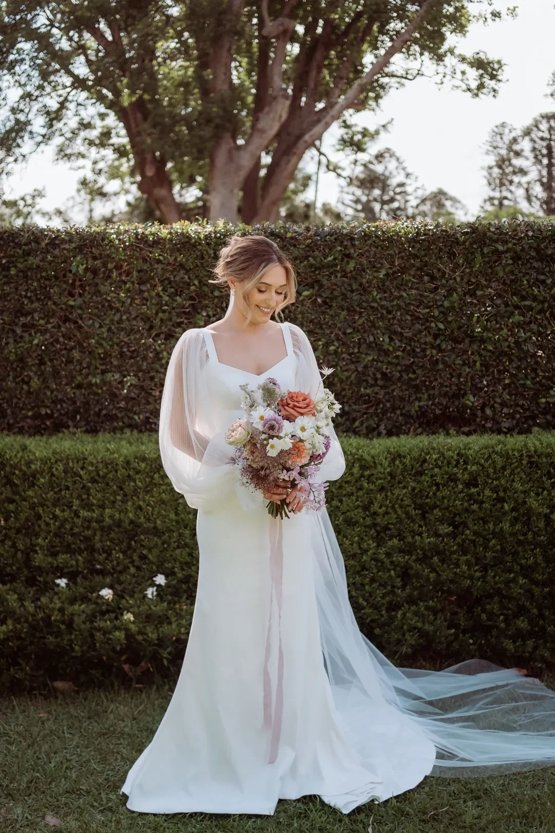 Bride Annalise holding her romantic garden-style bouquet in front of a manicured hedge at her wedding.