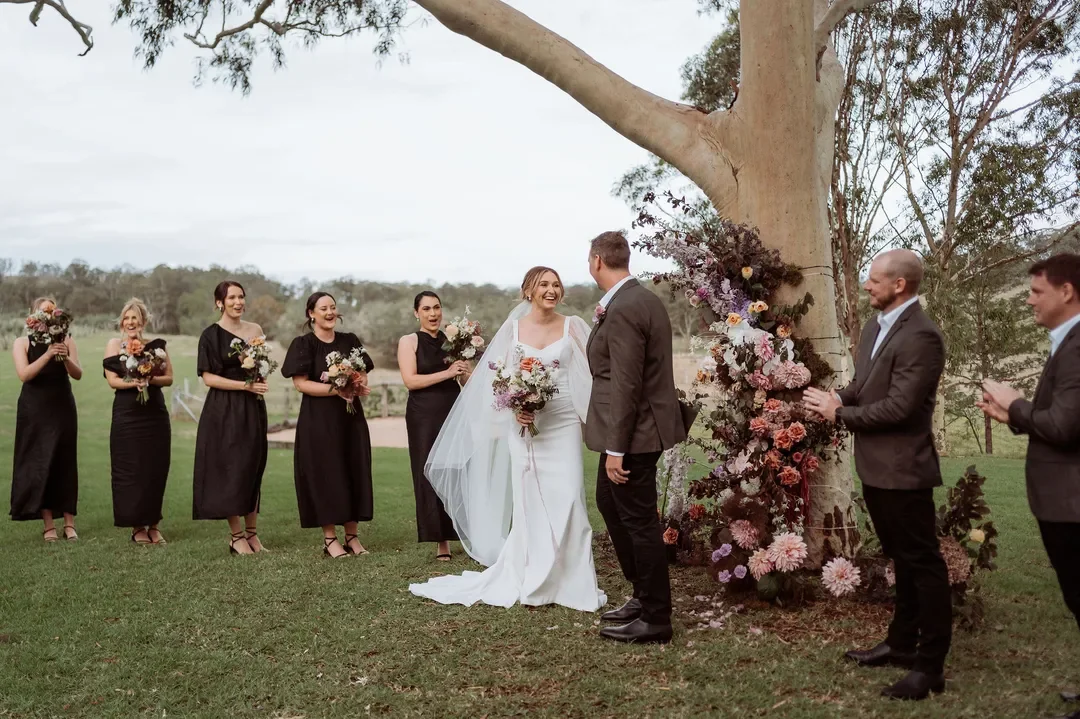 Bride Annalise and groom Mitchell smiling during their outdoor ceremony beneath a gum tree decorated with cascading florals, surrounded by their bridal party.