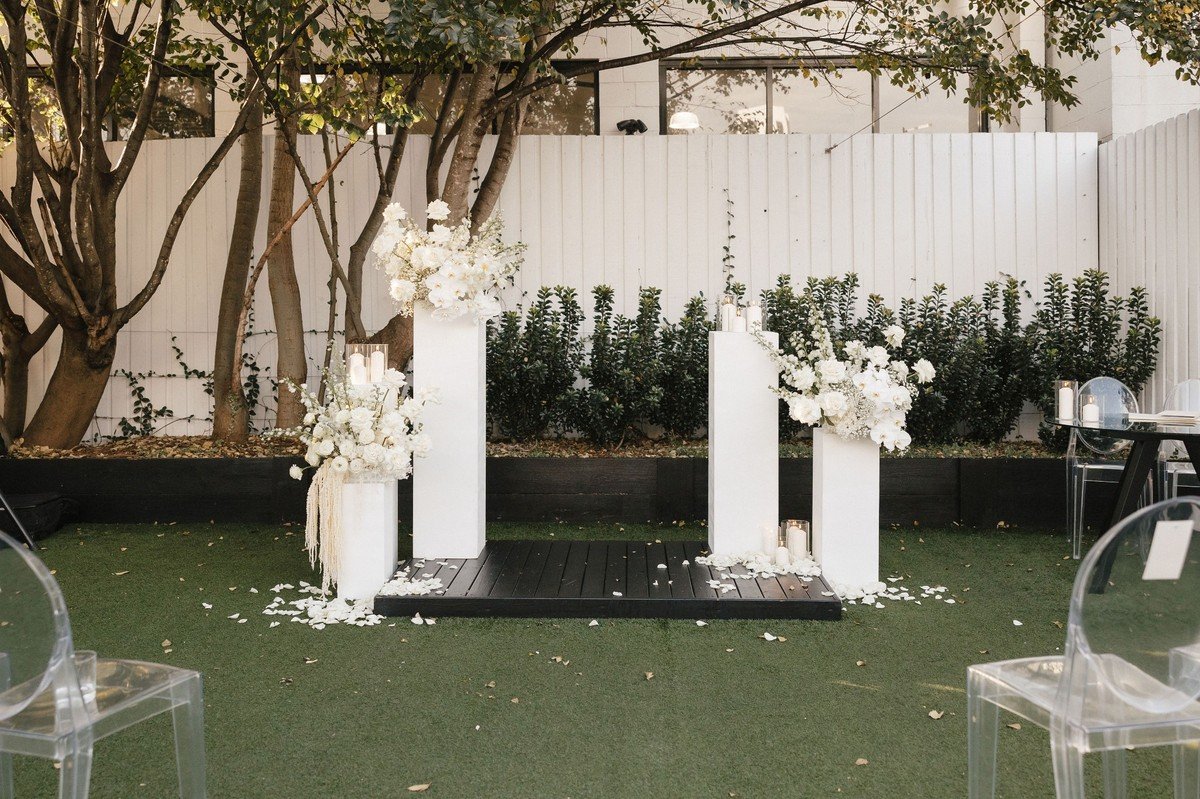 Minimal white ceremony floral pillars featuring roses, orchids, and candles set in a modern garden courtyard.