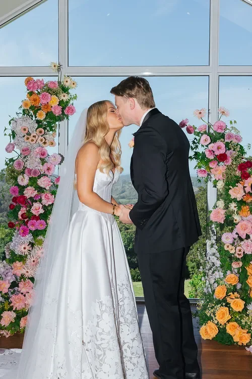 Bride and groom holding hands and kissing during their ceremony surrounded by lush floral installations