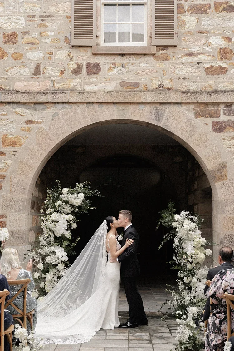 Bride and groom sharing their first kiss beneath a stone archway decorated with abundant white flowers.