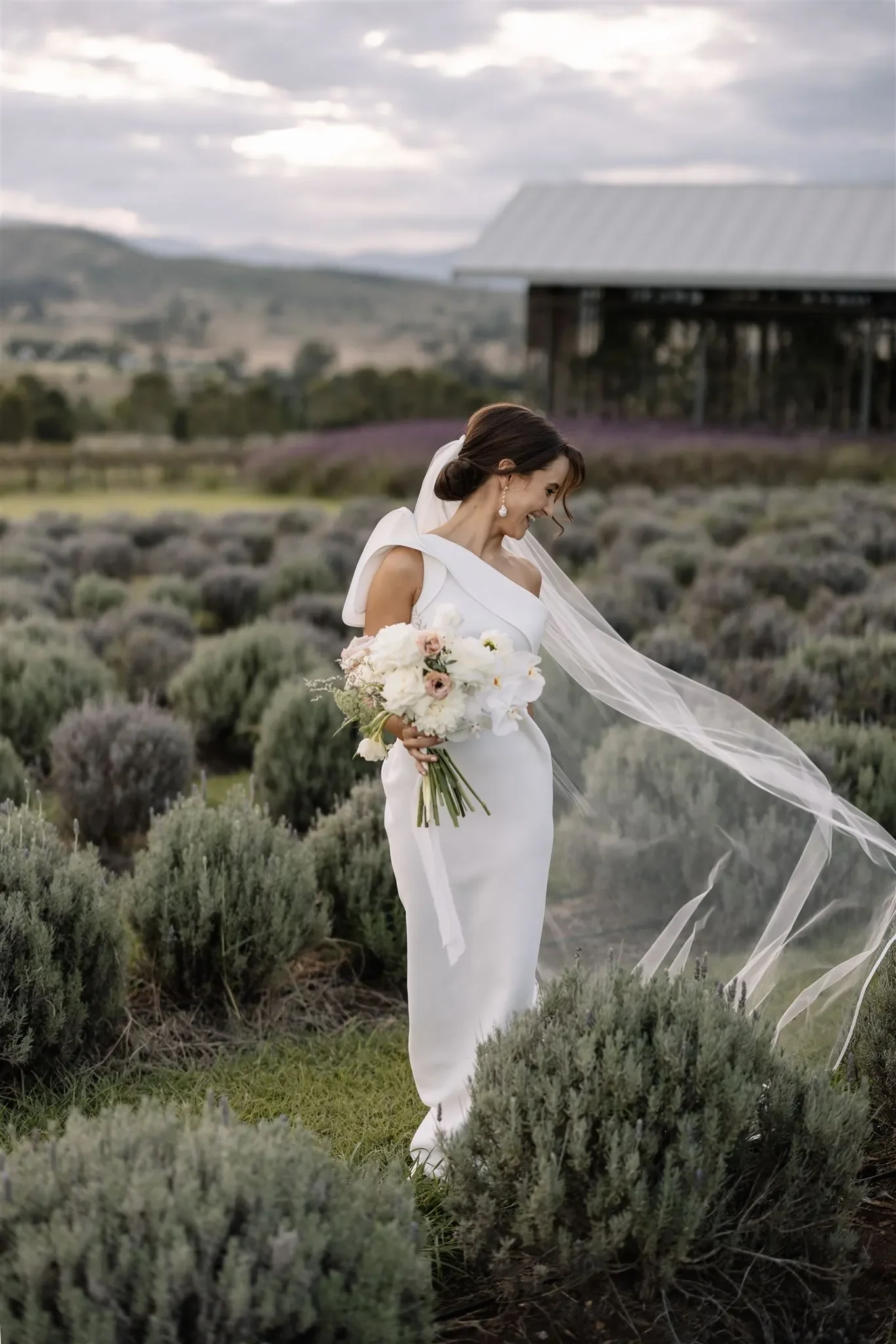 Bride holding a white floral bouquet during an outdoor garden wedding, wearing a modern long-sleeve gown with soft natural light filtering through trees