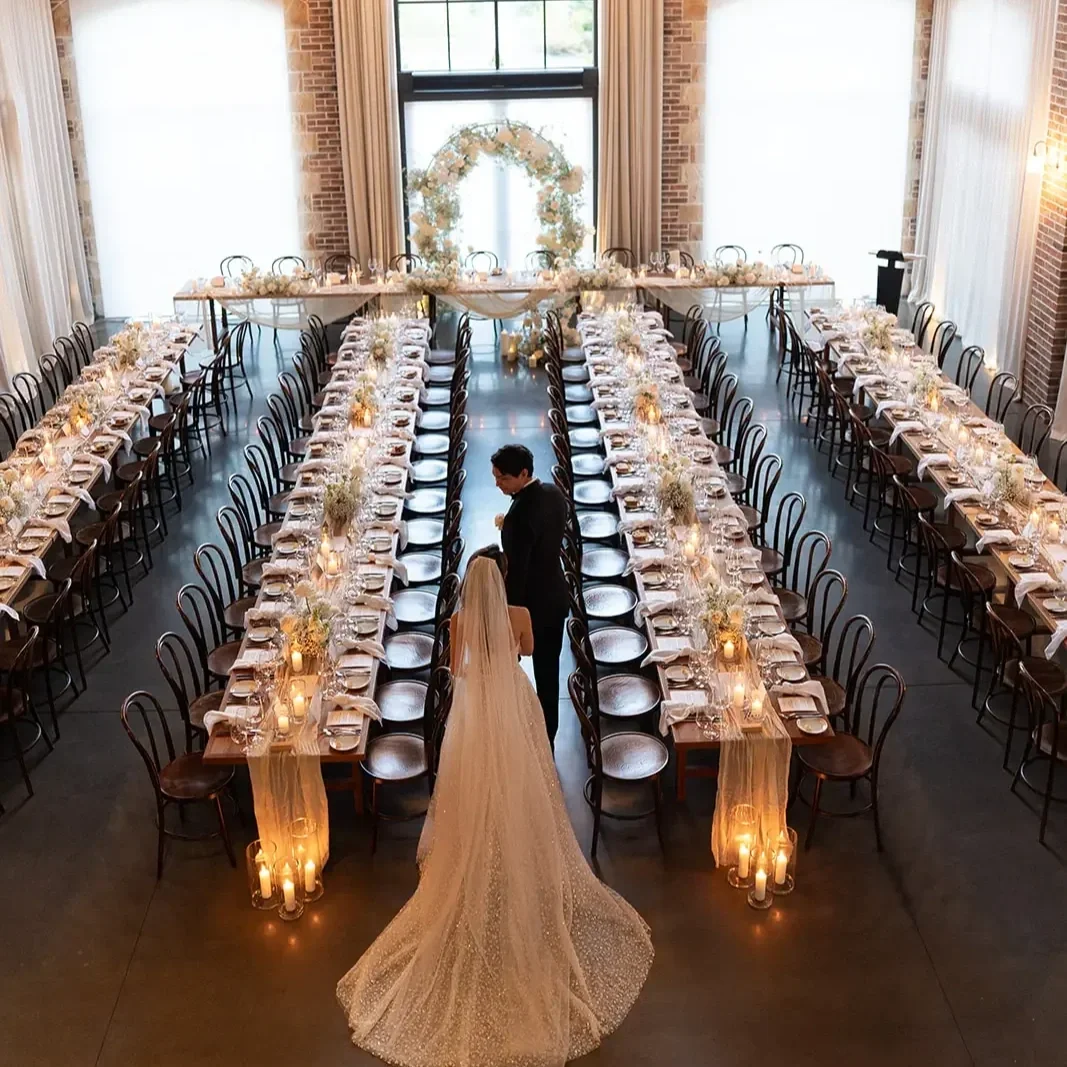 Long banquet-style wedding reception inside an elegant venue with chandeliers, candlelit tables and a soft white floral arbour at the head table