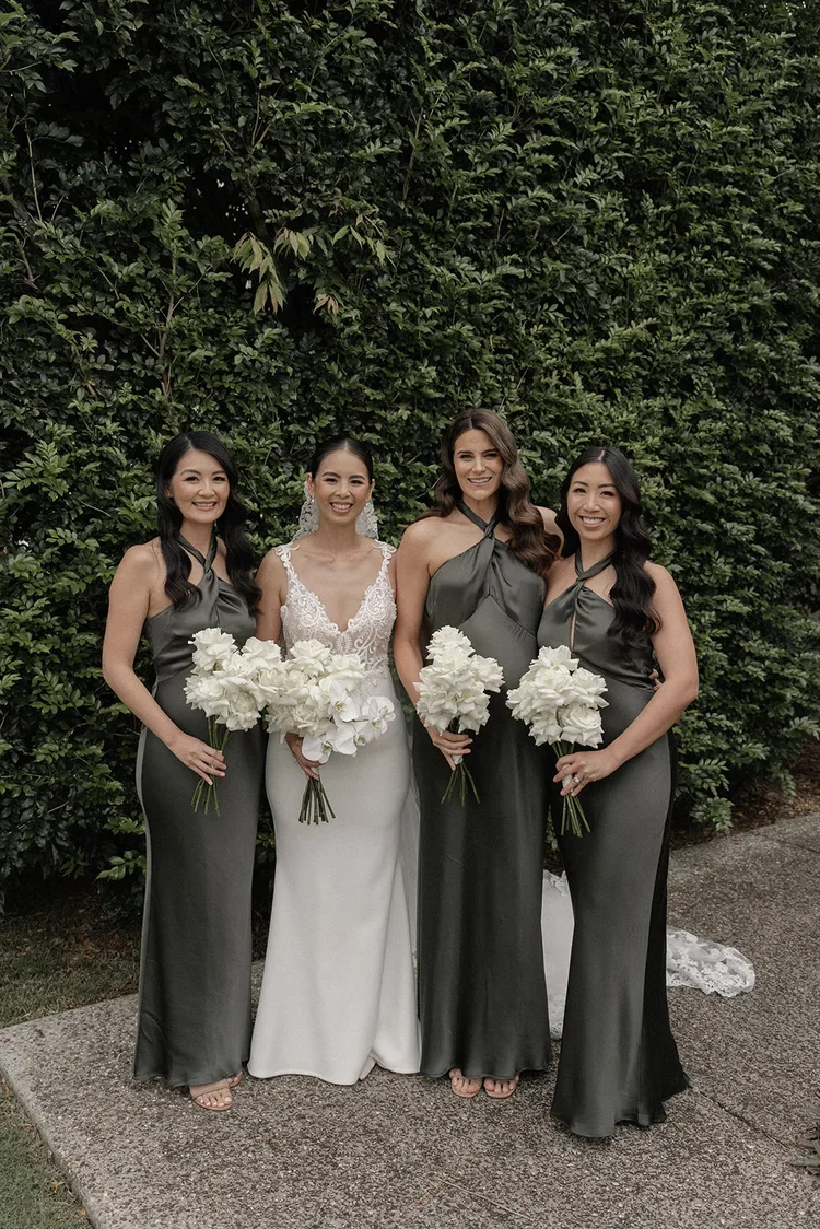 Bride and bridesmaids posing together outdoors, holding white bouquets, with bridesmaids wearing deep green satin dresses.