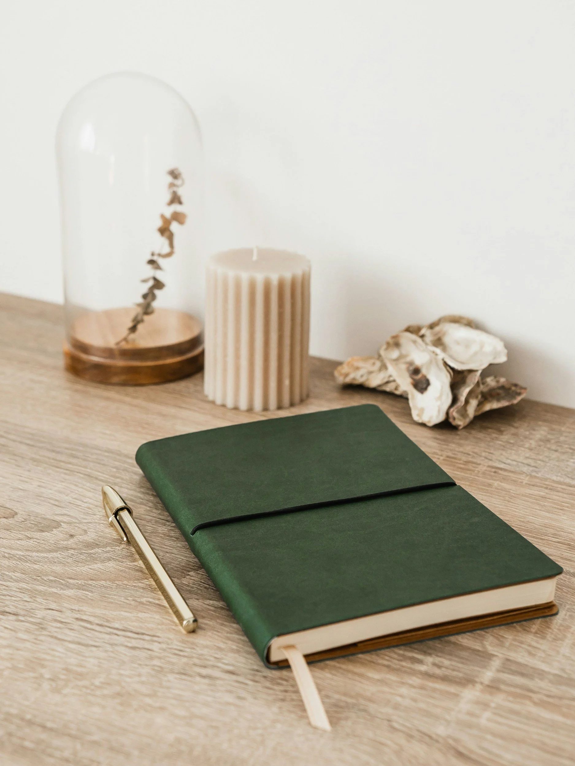 A green notebook with an elastic band, a gold pen, a beige candle, a glass dome with a wooden base covering a small object, and a cluster of seashells on a wooden surface.