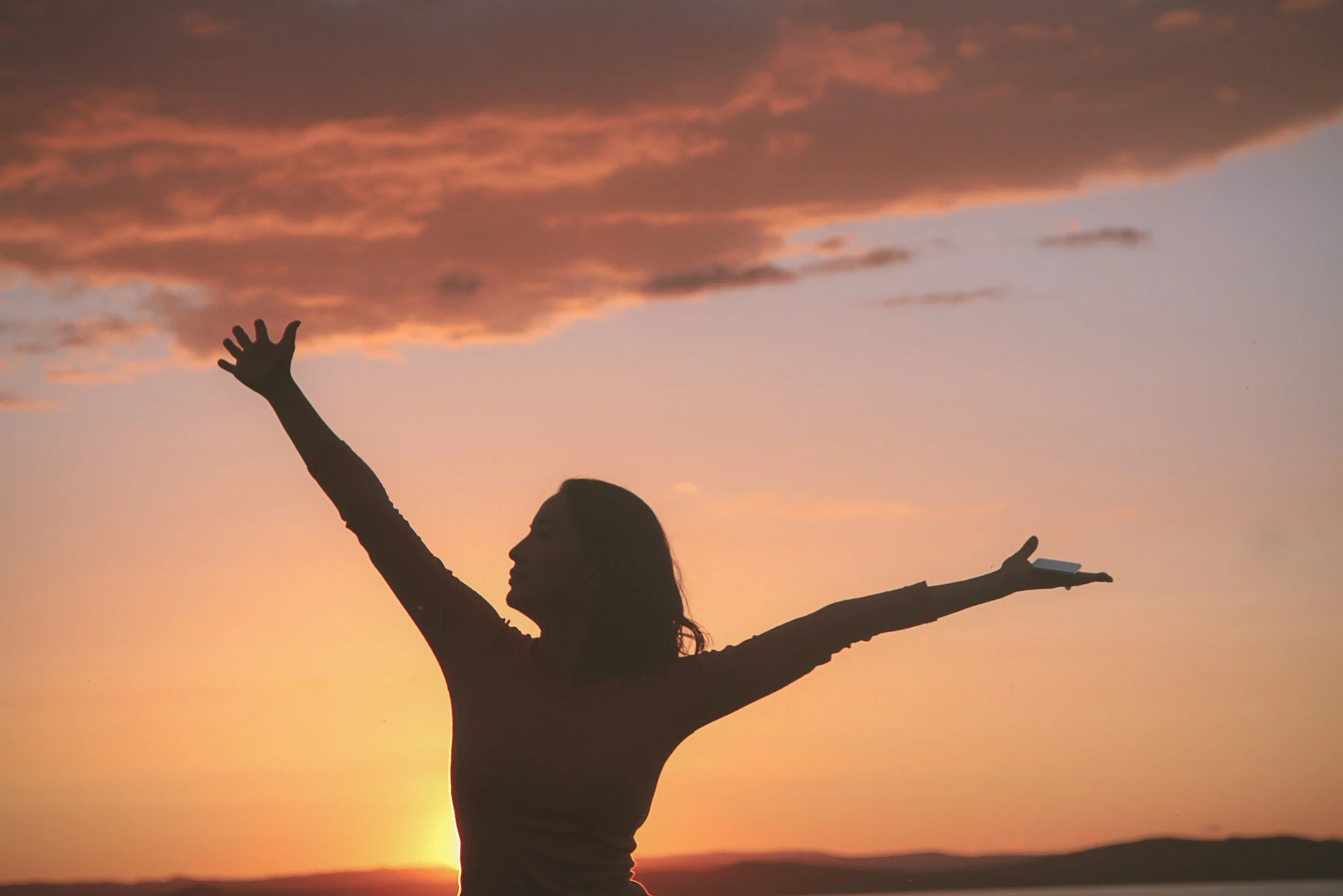 Silhouette of a woman standing outdoors at sunset, with her arms outstretched and looking up at the sky.