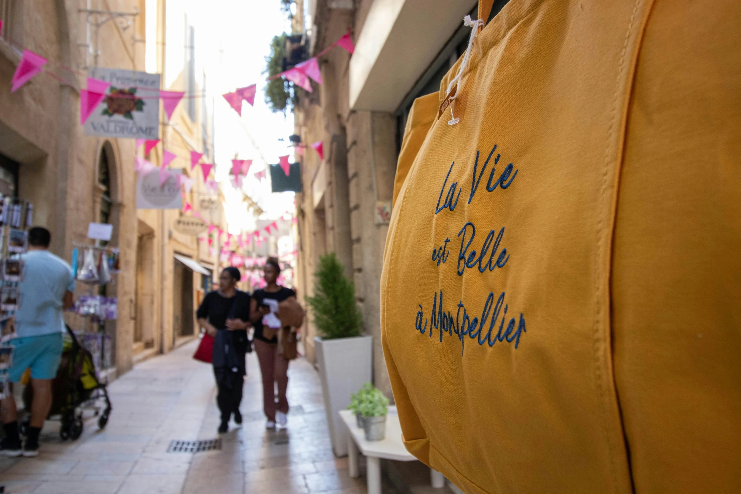 Street scene with people walking, pink bunting, and storefronts, with a yellow bag in the foreground.