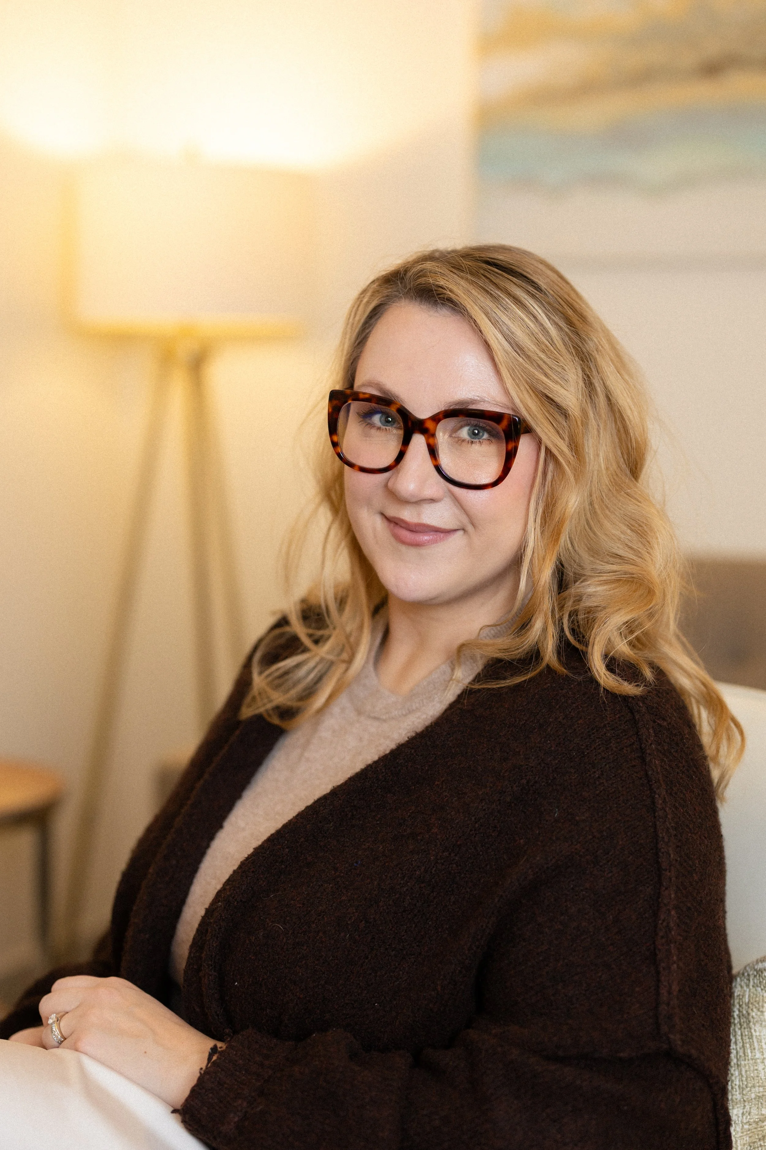 A woman with blonde hair, wearing glasses and a dark brown cardigan, sitting in a cozy room with a lamp and a painting in the background.