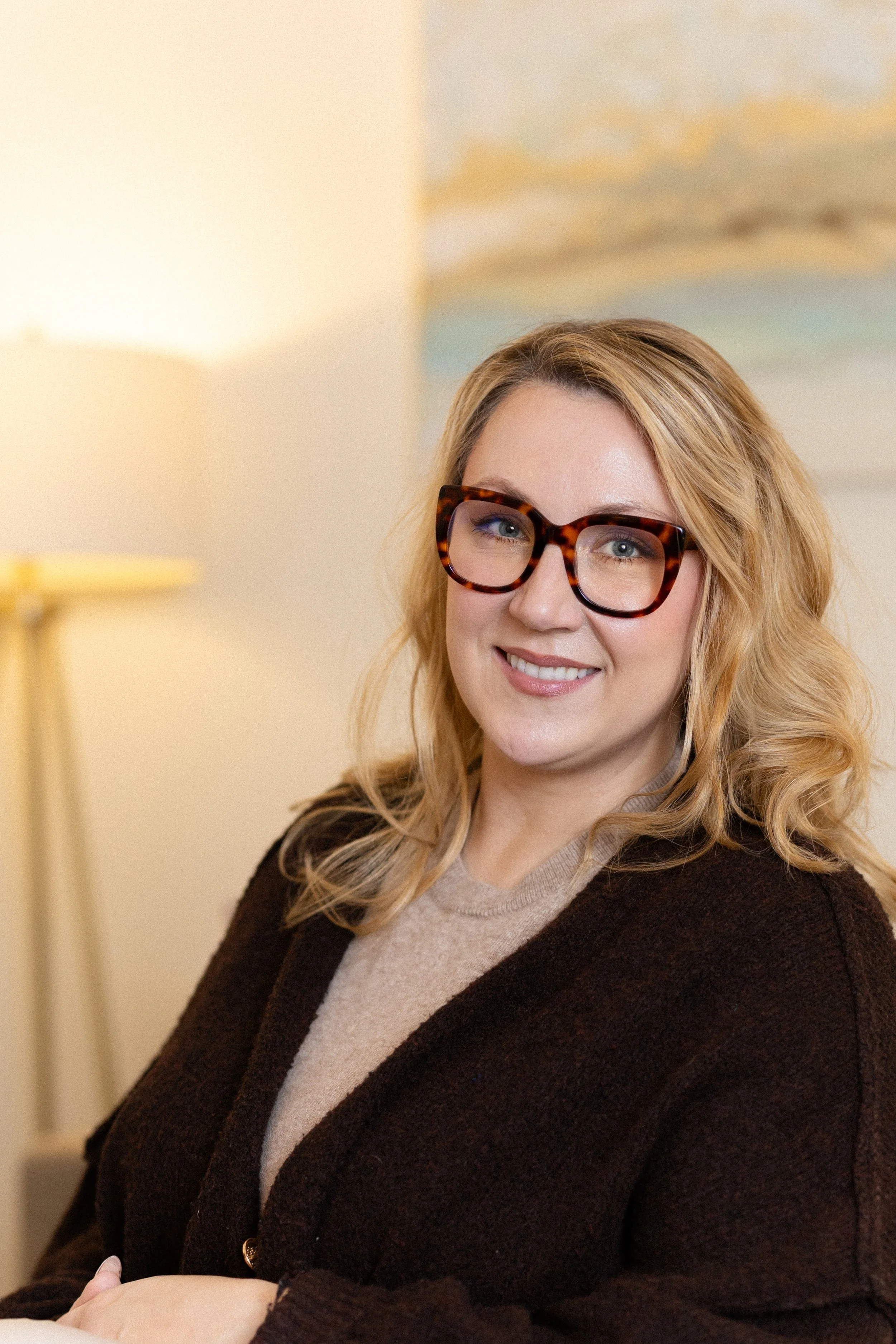 A woman with blonde curly hair, wearing tortoiseshell glasses and a brown cardigan, smiling indoors.