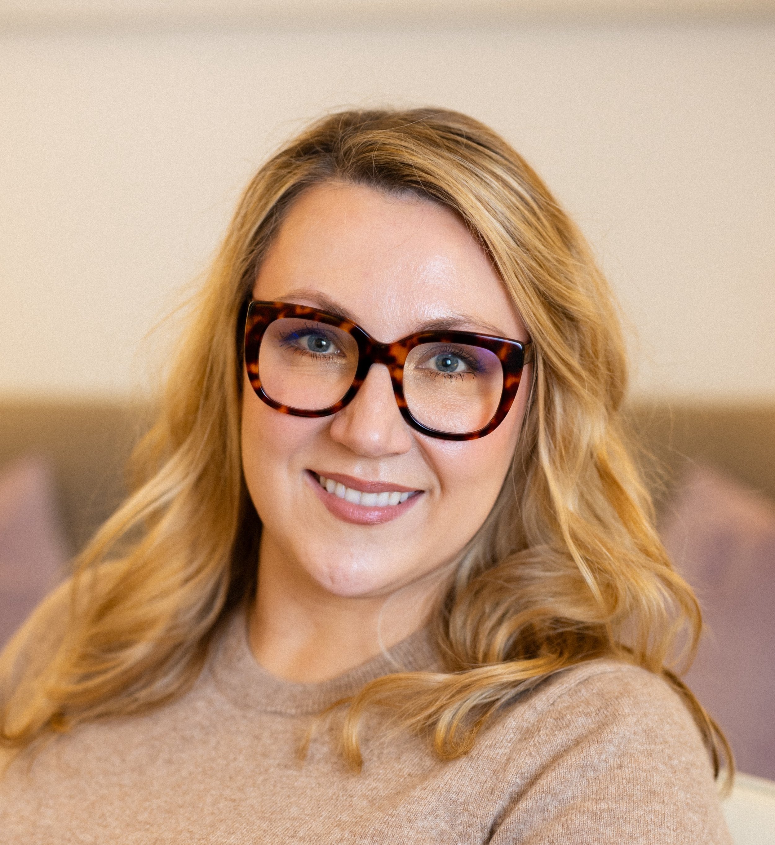 A smiling woman with blonde hair wearing tortoiseshell glasses and a beige sweater.