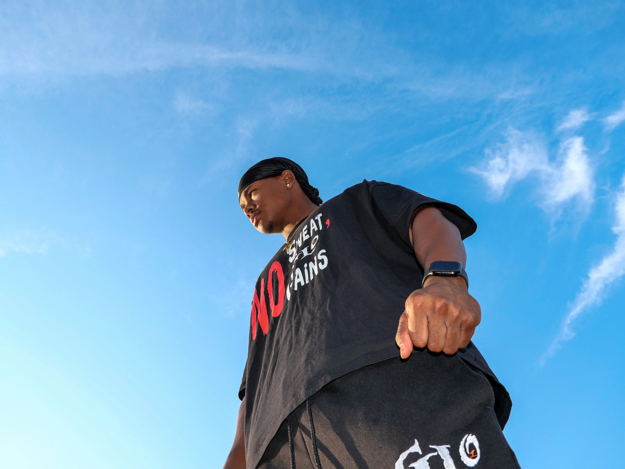 Close-up of a young man who is the gym content creator luckeylift wearing a black headscarf and a black T-shirt, looking to the side, with a blue sky and some clouds in the background.