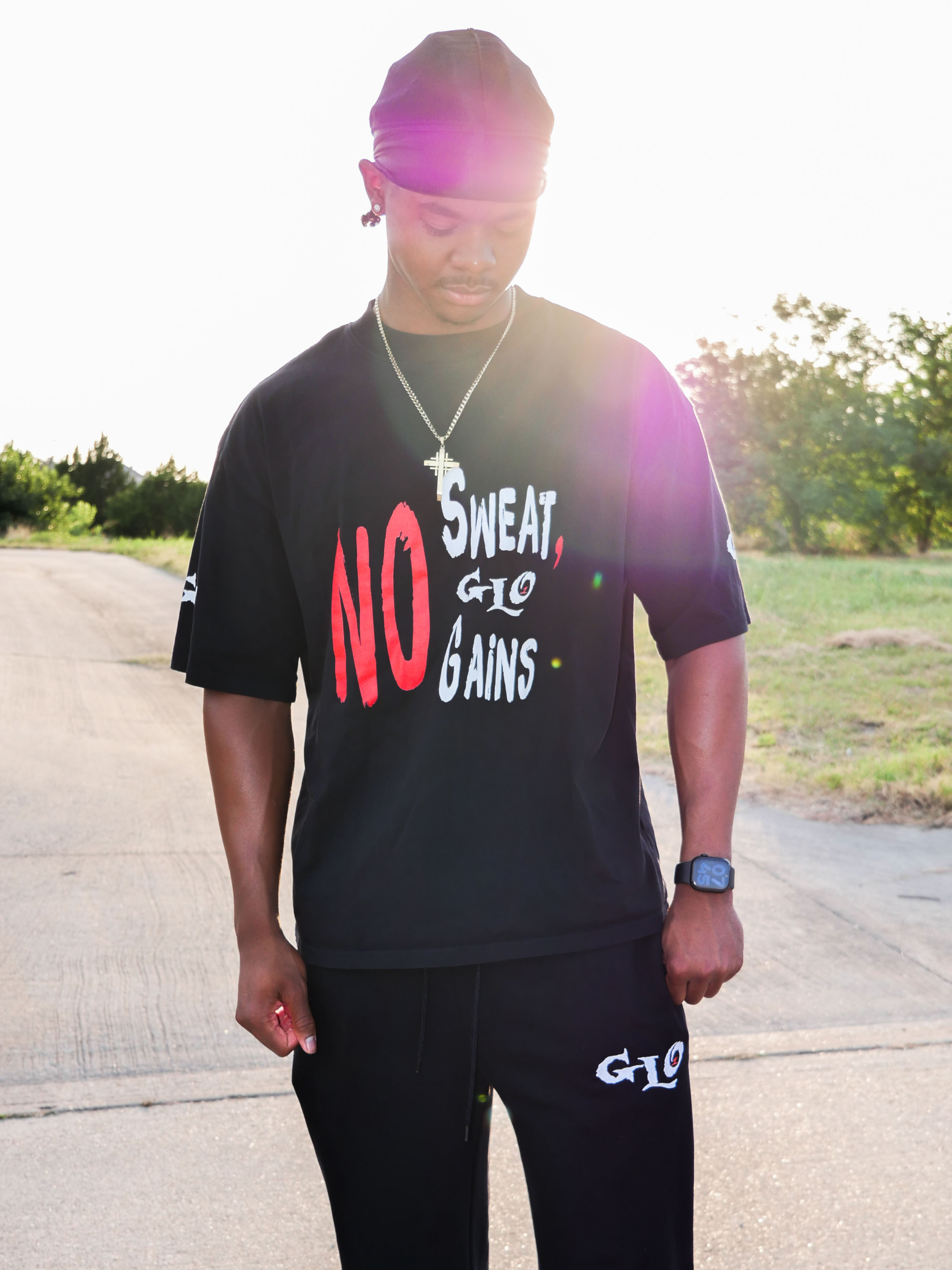 gym content creator named luckeylift standing outdoors wearing 1GLO t-shirt with white and red text, 1GLO black pants, a black headscarf, a silver chain with a cross pendant, a smartwatch, and earrings, with trees and a clear sky in the background.