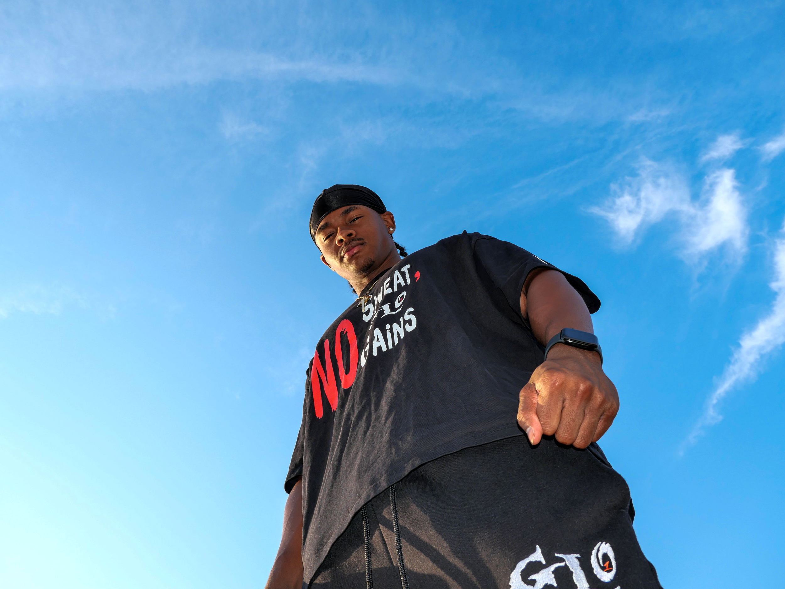 A young man who is the gym content creator luckeylift wearing a 1GLO black shirt , looking down at the camera against a bright blue sky with wispy clouds.