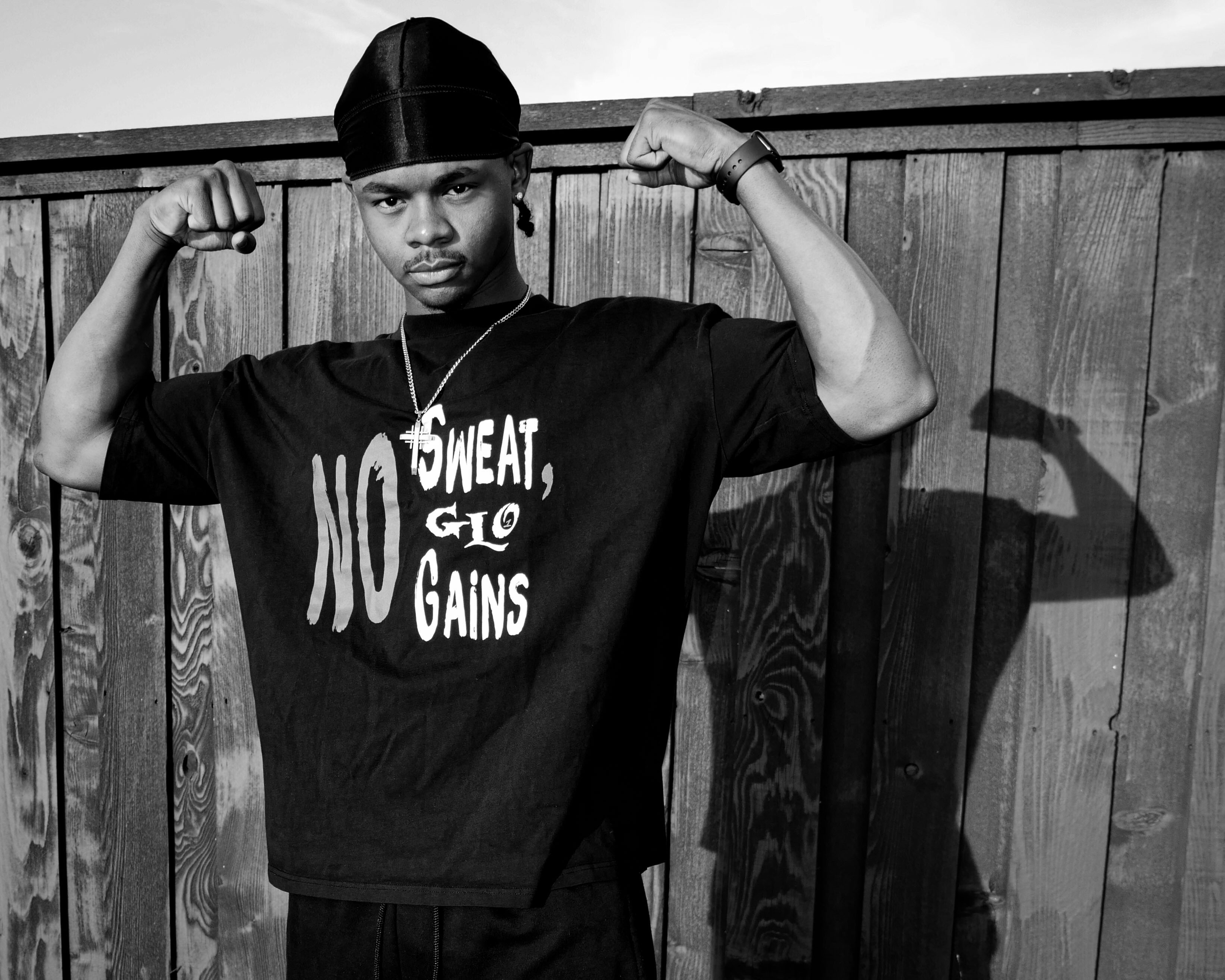 Gym content creator luckeylift flexing his biceps while looking confidently at the camera, wearing a beanie, jewelry, and a 1GLO T-shirt with words on it, standing in front of a wooden fence.