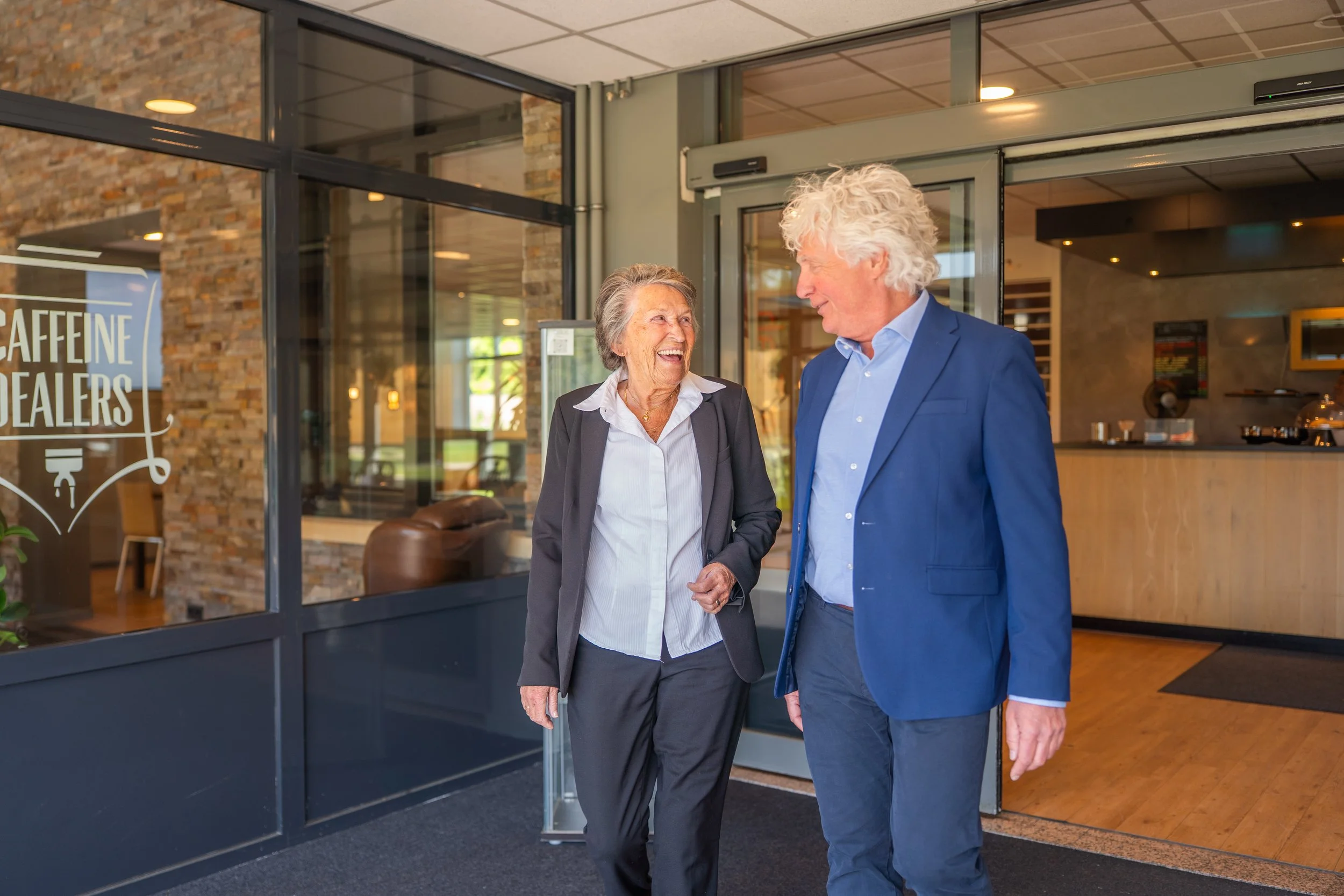 Een seniore man en vrouw, beide in formele kleding, lopen lachend door de hal van een bedrijfspand.