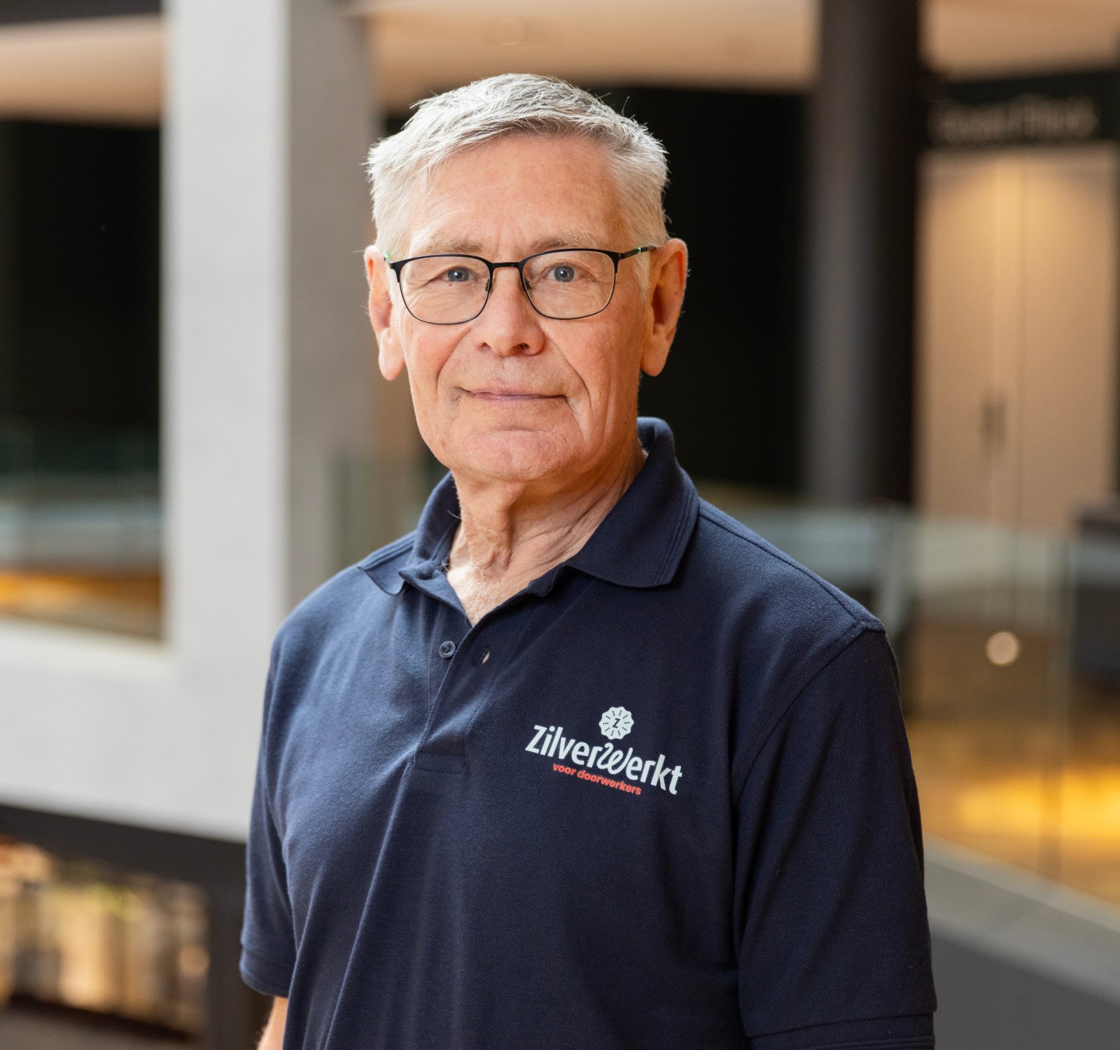 Portret van een seniore man met kort wit haar, zwarte bril en donkerblauwe ZilverWerkt-polo.