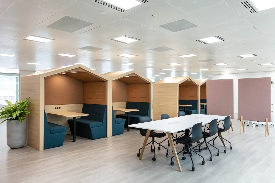 Un espace de bureau moderne avec des cabines en bois en forme de maison, des tables blanches et des chaises noires, avec des panneaux séparateurs et une grande plante en pot.
