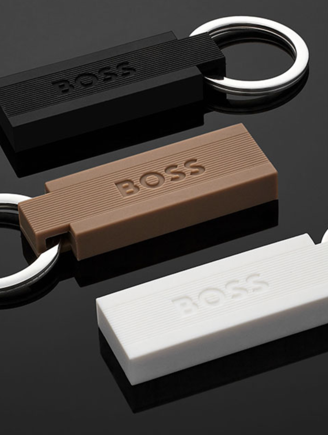 Porte-clés rectangulaires avec le mot 'BOSS' en noir, marron et blanc sur fond sombre. Anneau métallique attaché à chaque porte-clés.