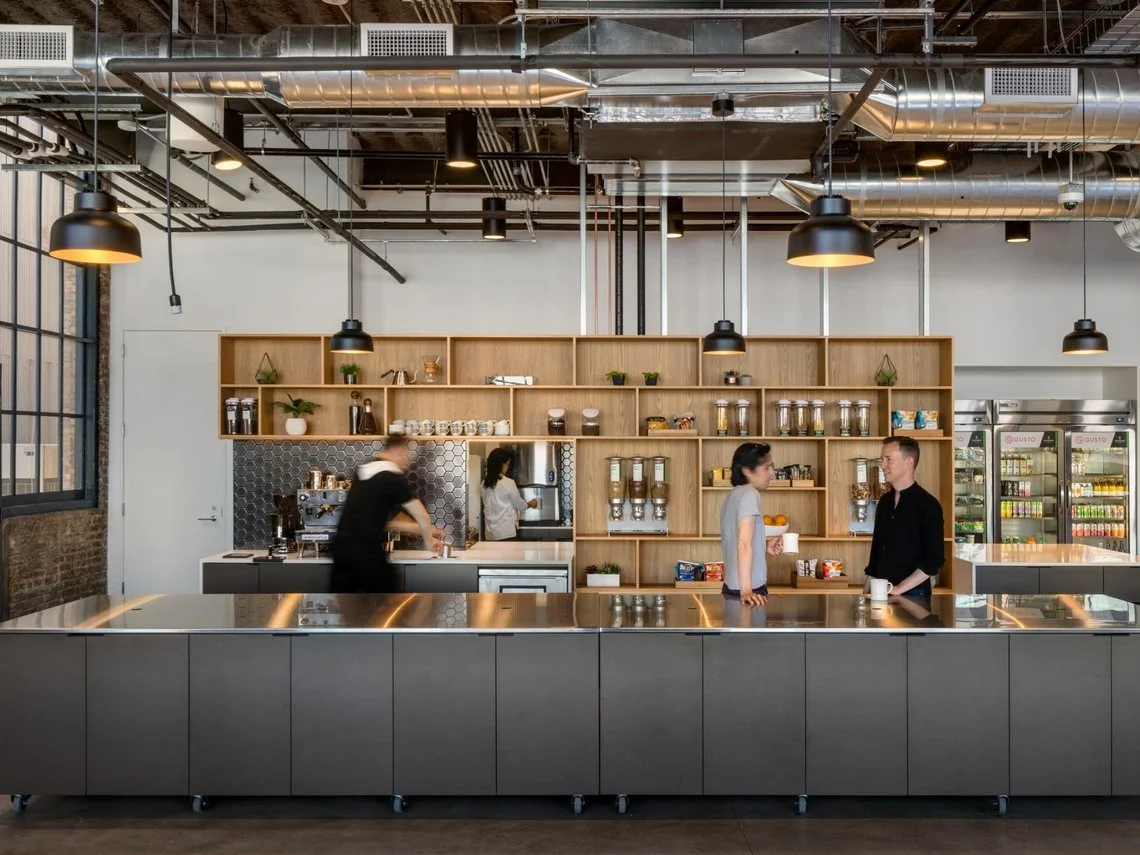 Cuisine industrielle moderne avec des étagères en bois, des cafetières, des rangements de nourriture. Trois personnes interagissent, l'une prépare une boisson. Éclairage industriel au plafond."