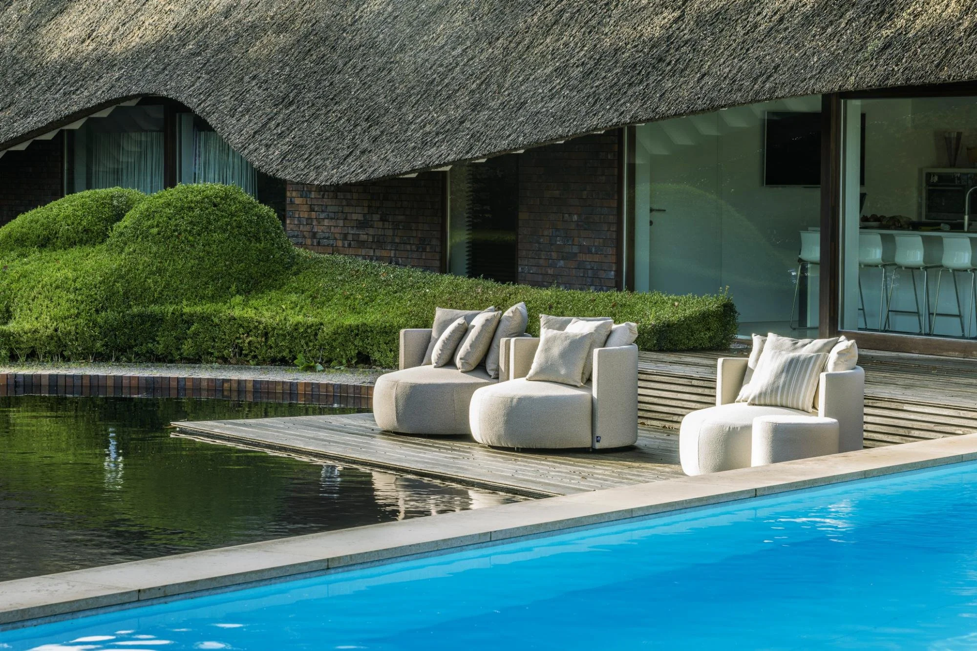 Façade moderne d'une maison avec un mobilier extérieur près de la piscine, comprenant des fauteuils et des coussins, entourée de buissons verts et avec des vitres donnant sur une cuisine intérieure.