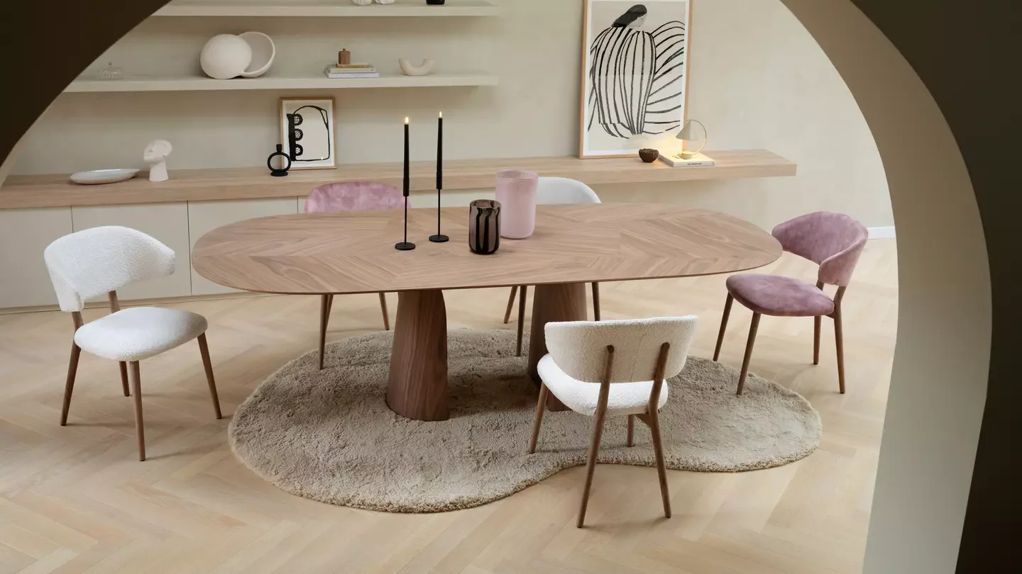 Salle à manger avec une table en bois, entourée de cinq chaises en tissu rose et blanc, sur un tapis beige. Décor moderne et minimaliste avec étagères murales contenant des objets décoratifs, une lampe, des cadres d'art et des vases.