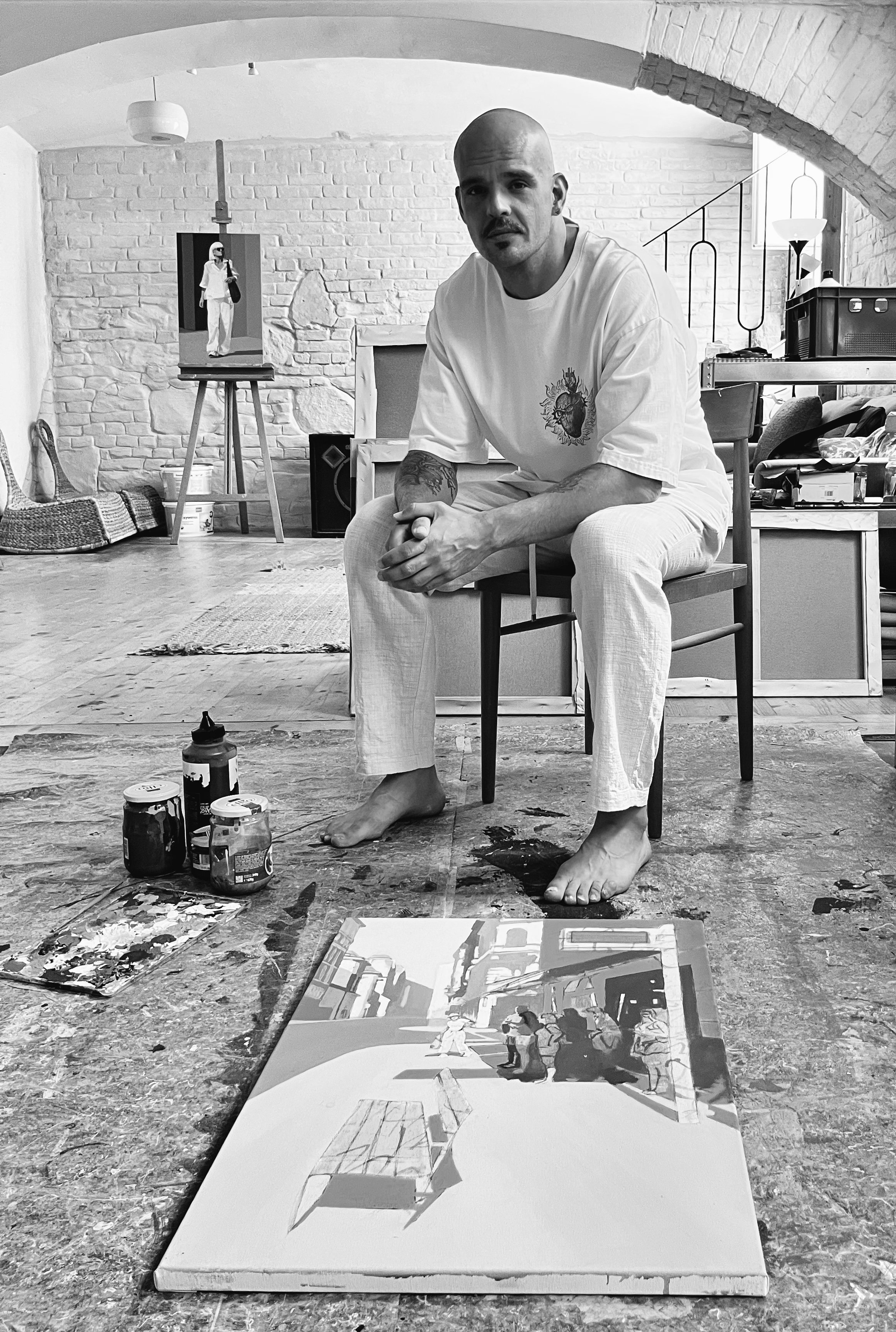 An artist, Roman Rieth, in a studio sitting on a chair, surrounded by art supplies and paintings, with a brick wall and an artwork of a woman on an easel in the background.