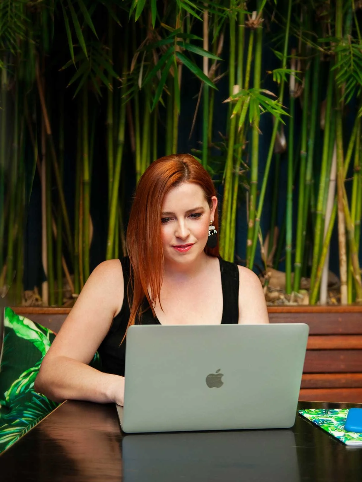 Kate Wilson - a woman with red hair working on a laptop at a wooden table, surrounded by green tropical plants in a cozy indoor setting.