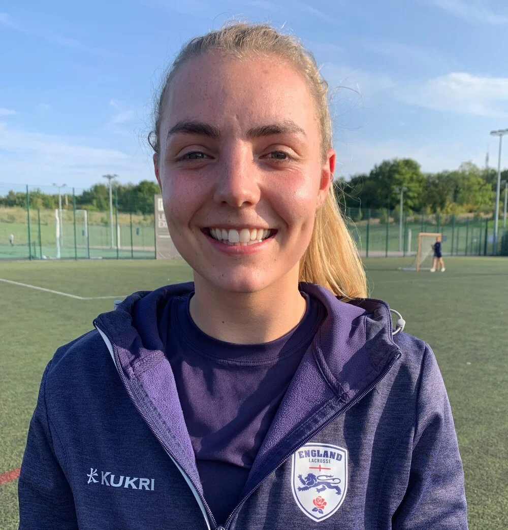Young woman smiling on a sports field wearing an England lacrosse jacket.