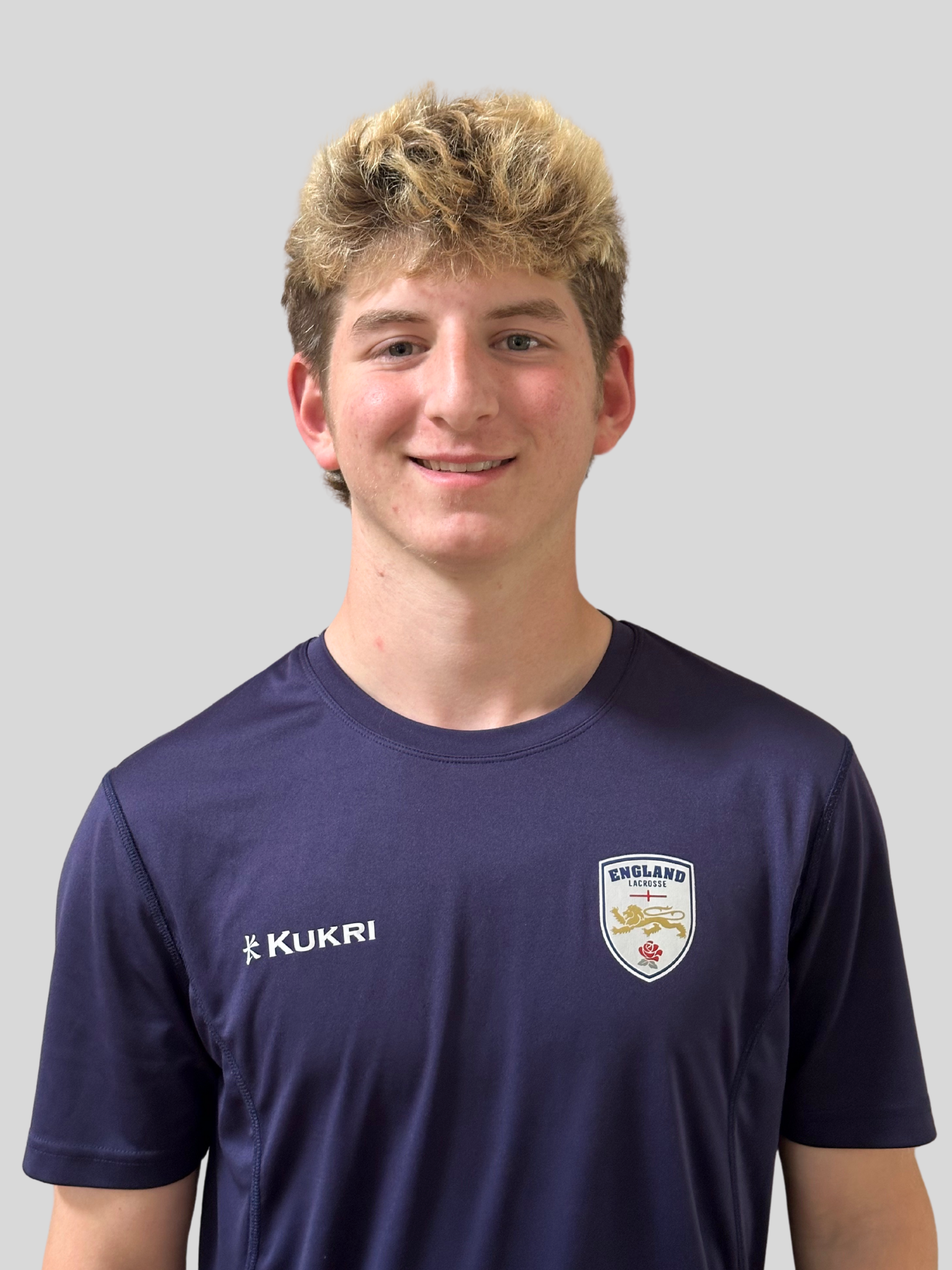 A young man with brown curly hair wearing a navy sports jacket standing in front of a large English flag with a textured, worn look.