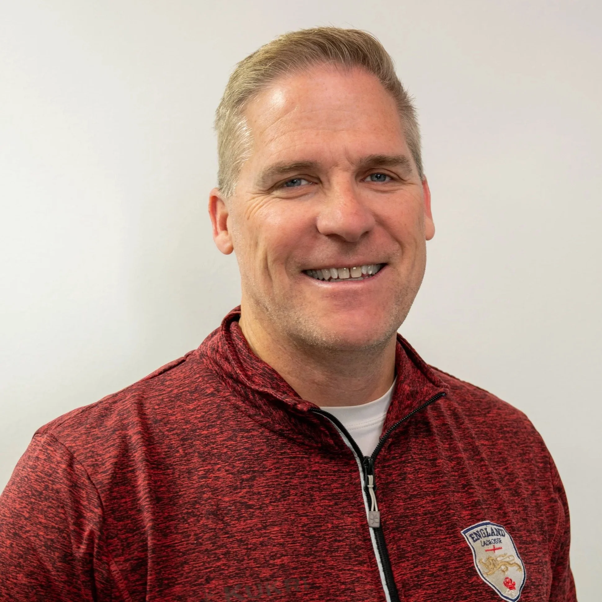 Man in a red zip-up sweater with an 'England Lacrosse' emblem on a neutral background.