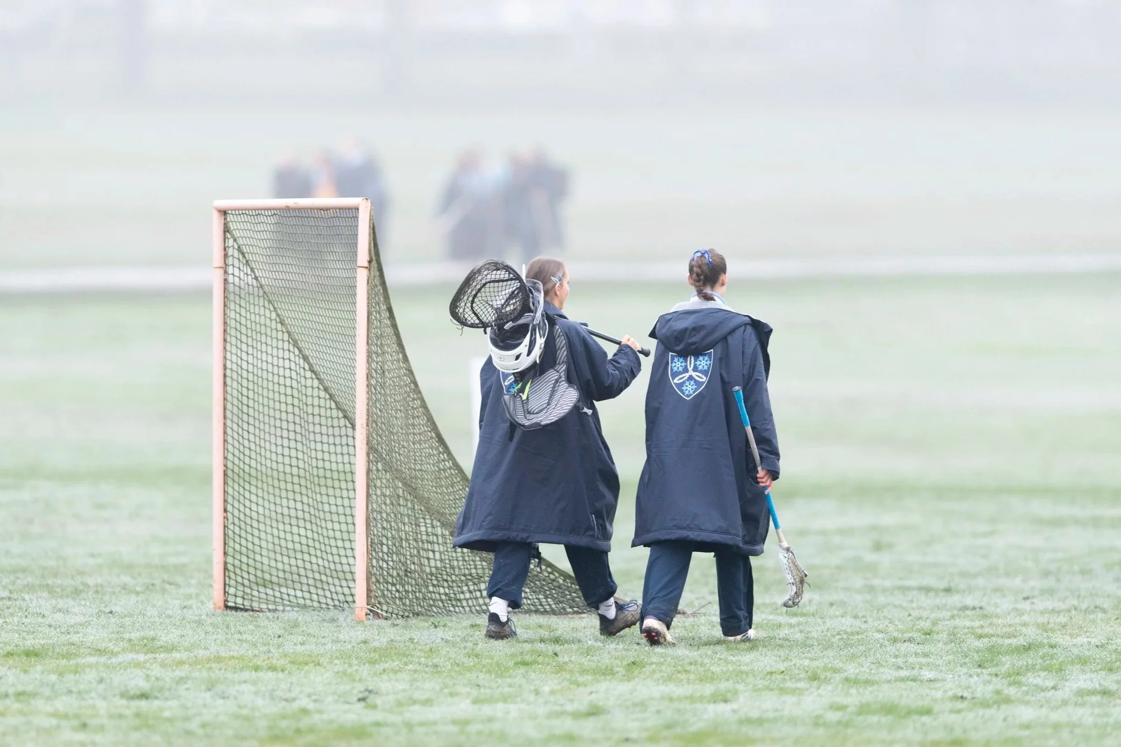 Two players walk across field with sticks in hands, next to a goal