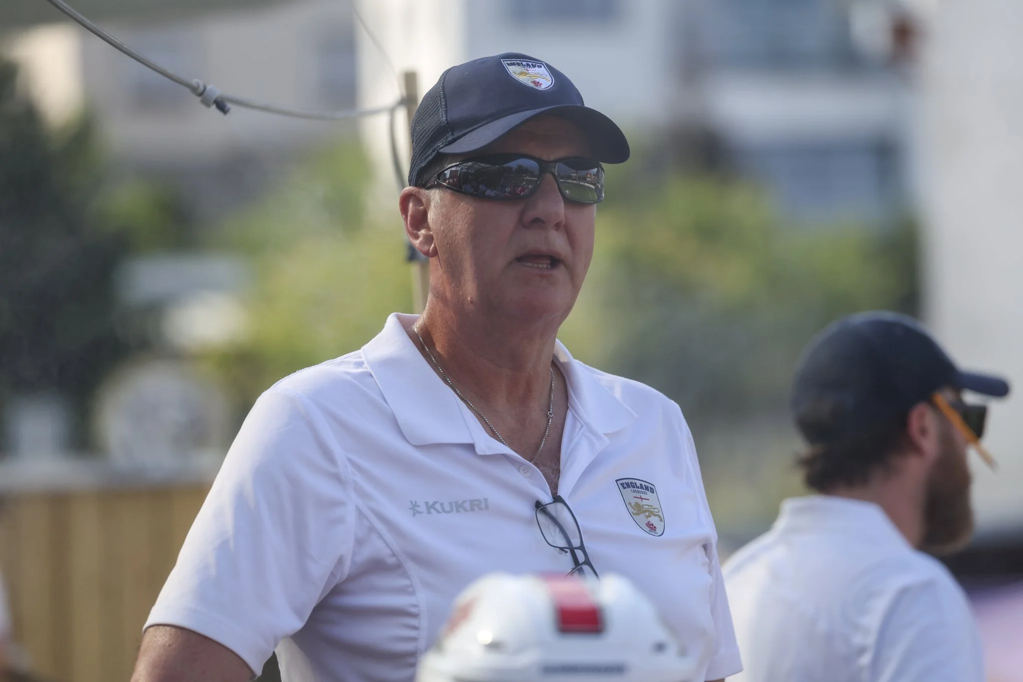 Walt Christianson looks off camera wearing sunglasses and an England Lacrosse cap and polo shirt