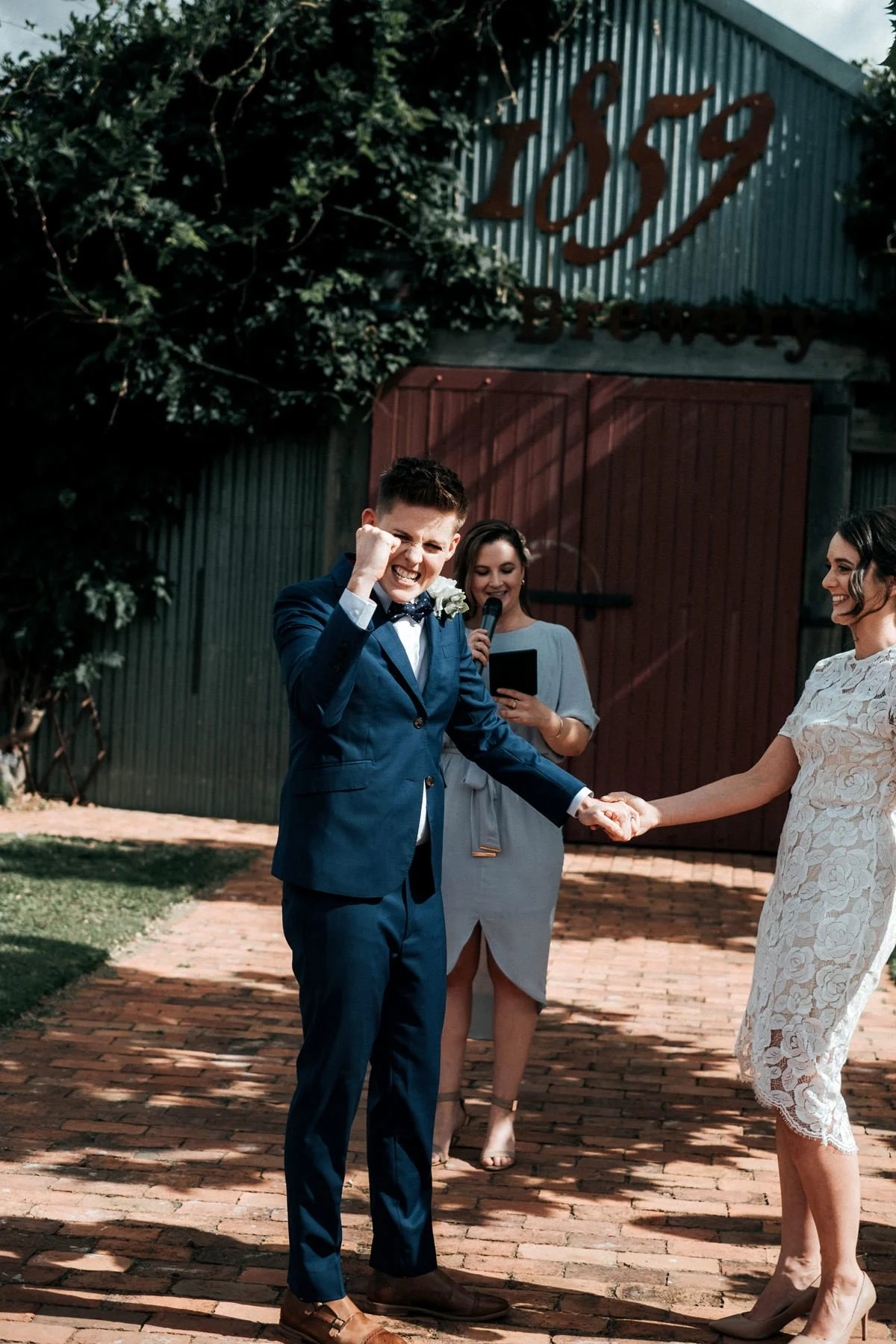 One bride cheering, holding her brides hand as the celebrant Married By Leah behind them announces them married at the Beekeepers Inn, Bathurst