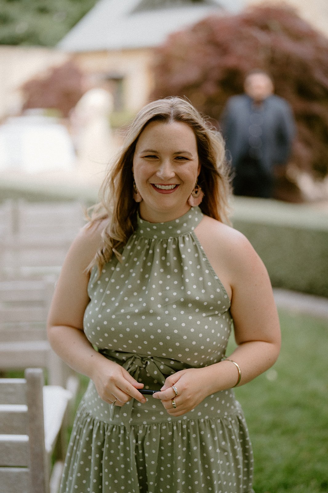Smiling Married By Leah in green polka dot dress standing outdoors at a daytime event, with blurred background and other people.