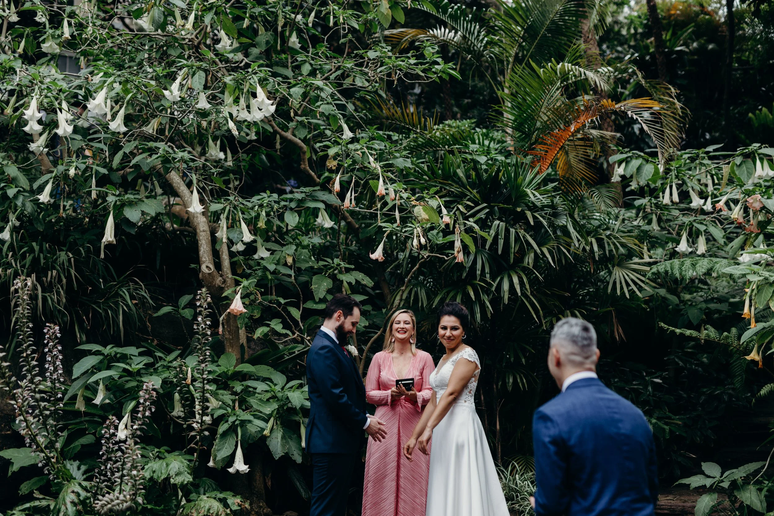 A bride and groom about to exchange rings at a wedding ceremony in Wendy's Secret Garden, Lavender Bay in front of a large green leaves and white trumpet-shaped flowers in the background. Celebrant Married By Leah in between the bride and groom.