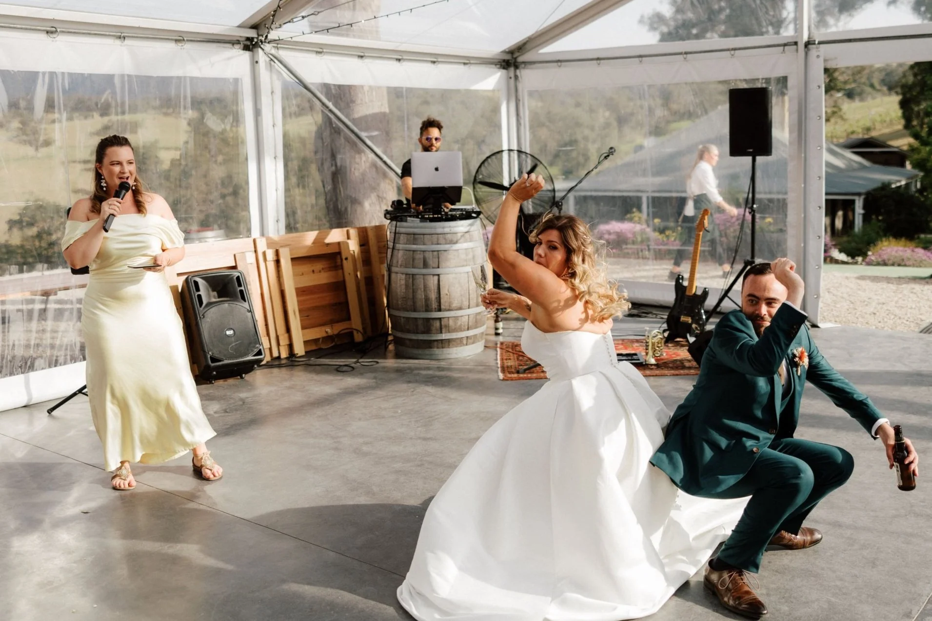 Bride in white wedding dress and groom in dark suit dancing at wedding reception under a tent, with Married By Leah in a yellow dress MCing the reception and a DJ in the background, outdoor setting with trees and tents visible outside.