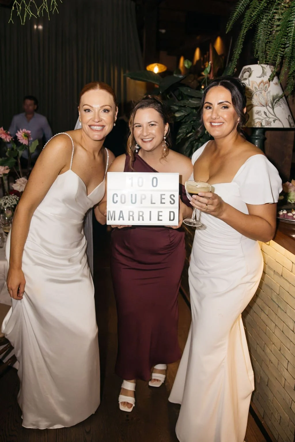 Two brides with Celebrant Married By Leah holding a "100 couples married" sign in the middle of them at the Grounds of Alexandria during their reception.