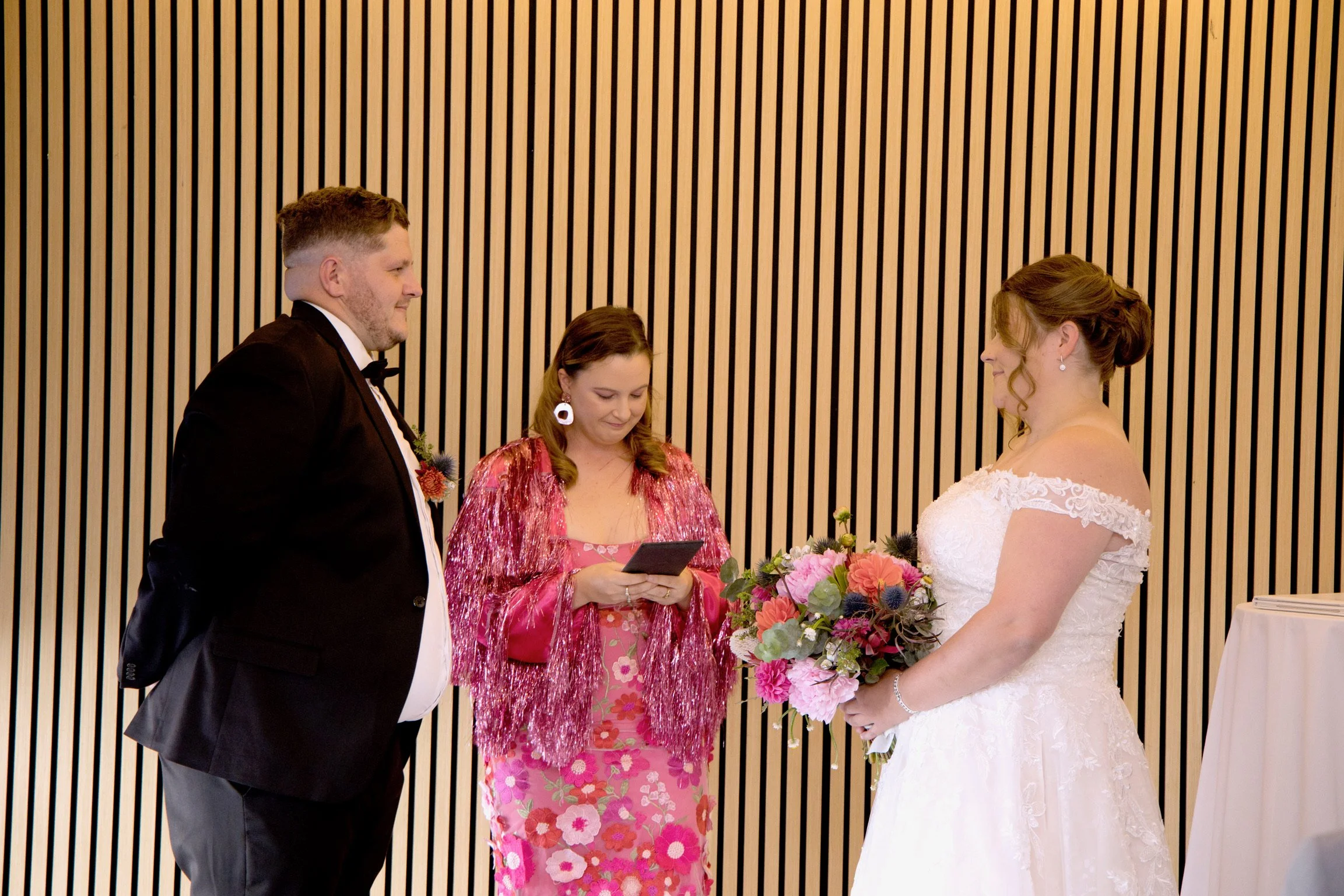 Groom and Bride in the middle of their ceremony with their celebrant Married By Leah in the middle in pink sparkly jacket